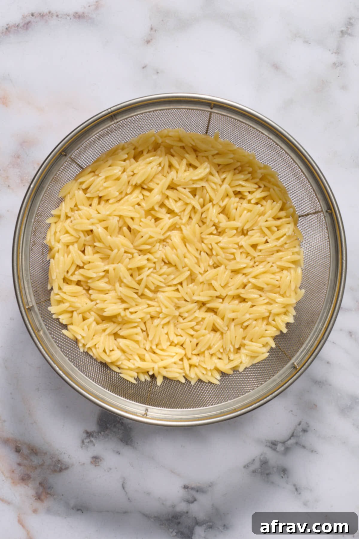 Cooked orzo pasta, perfectly al dente, being drained in a colander under cold running water.