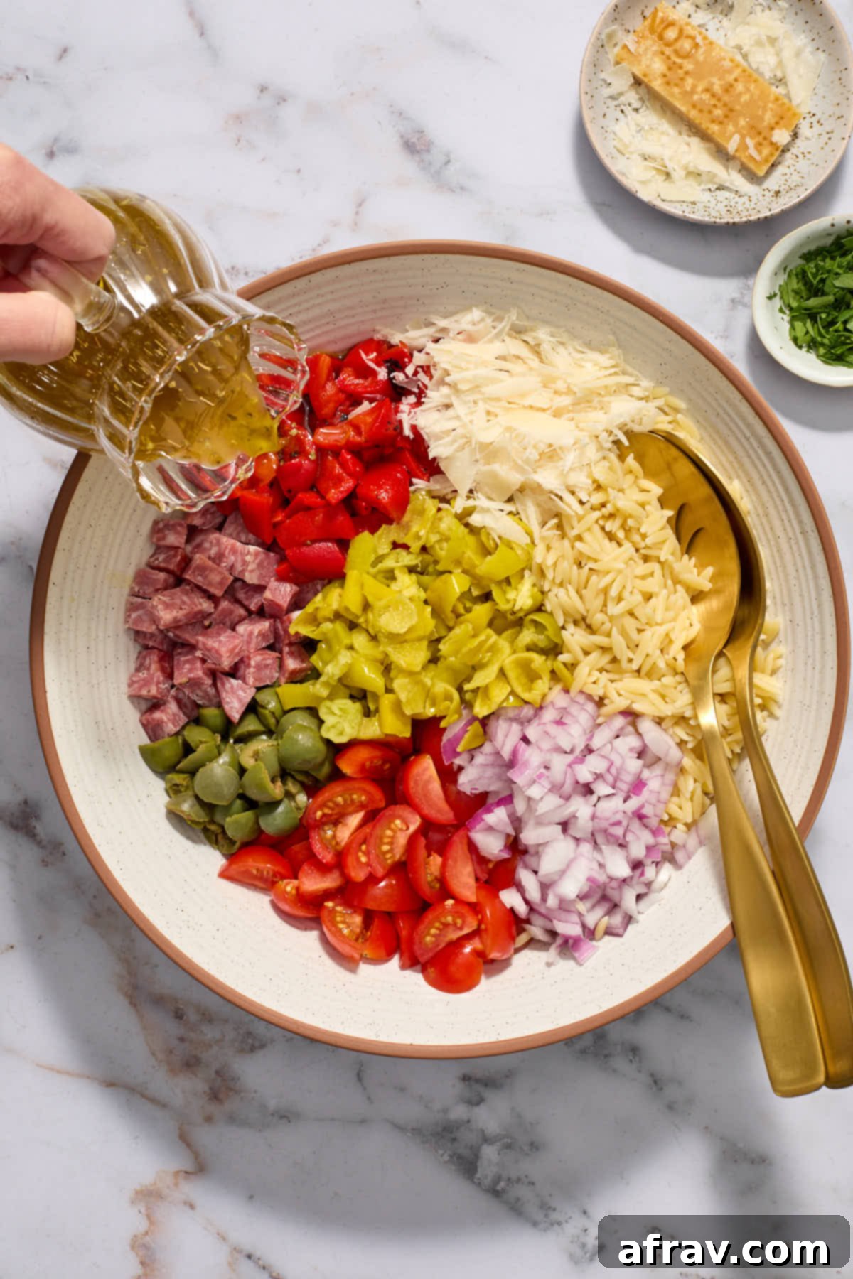 Prepared orzo pasta and salad ingredients arranged in a large white bowl, with homemade Italian dressing being poured over them.