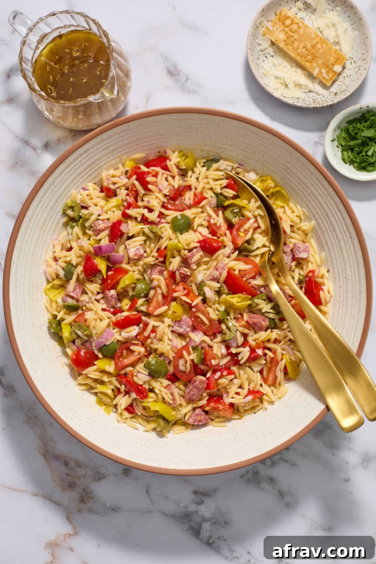 A large white bowl filled with Italian Orzo Salad, with gold salad servers tucked into the side, showcasing the colorful ingredients.