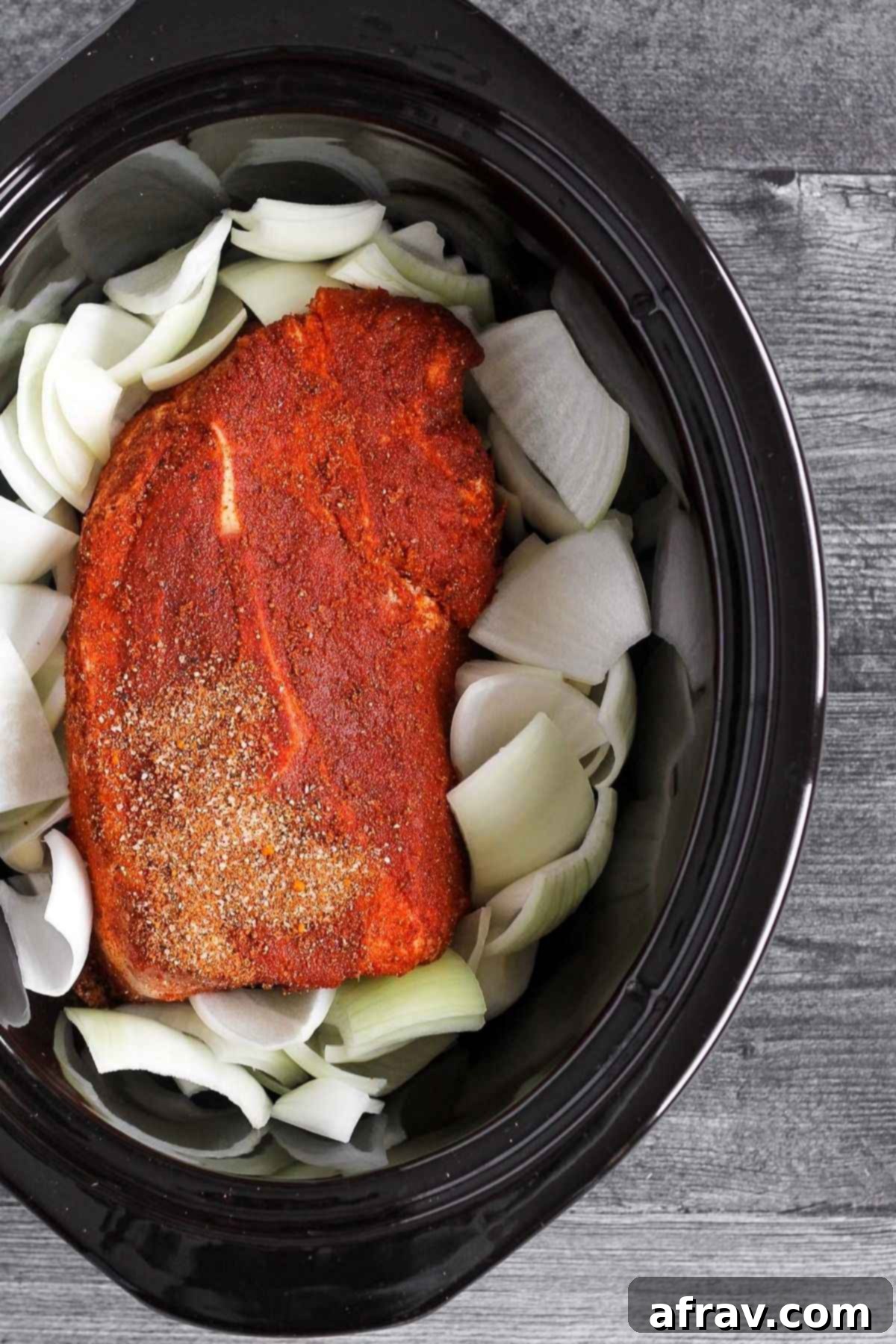 Raw pork with rub and raw onions in a slow cooker.