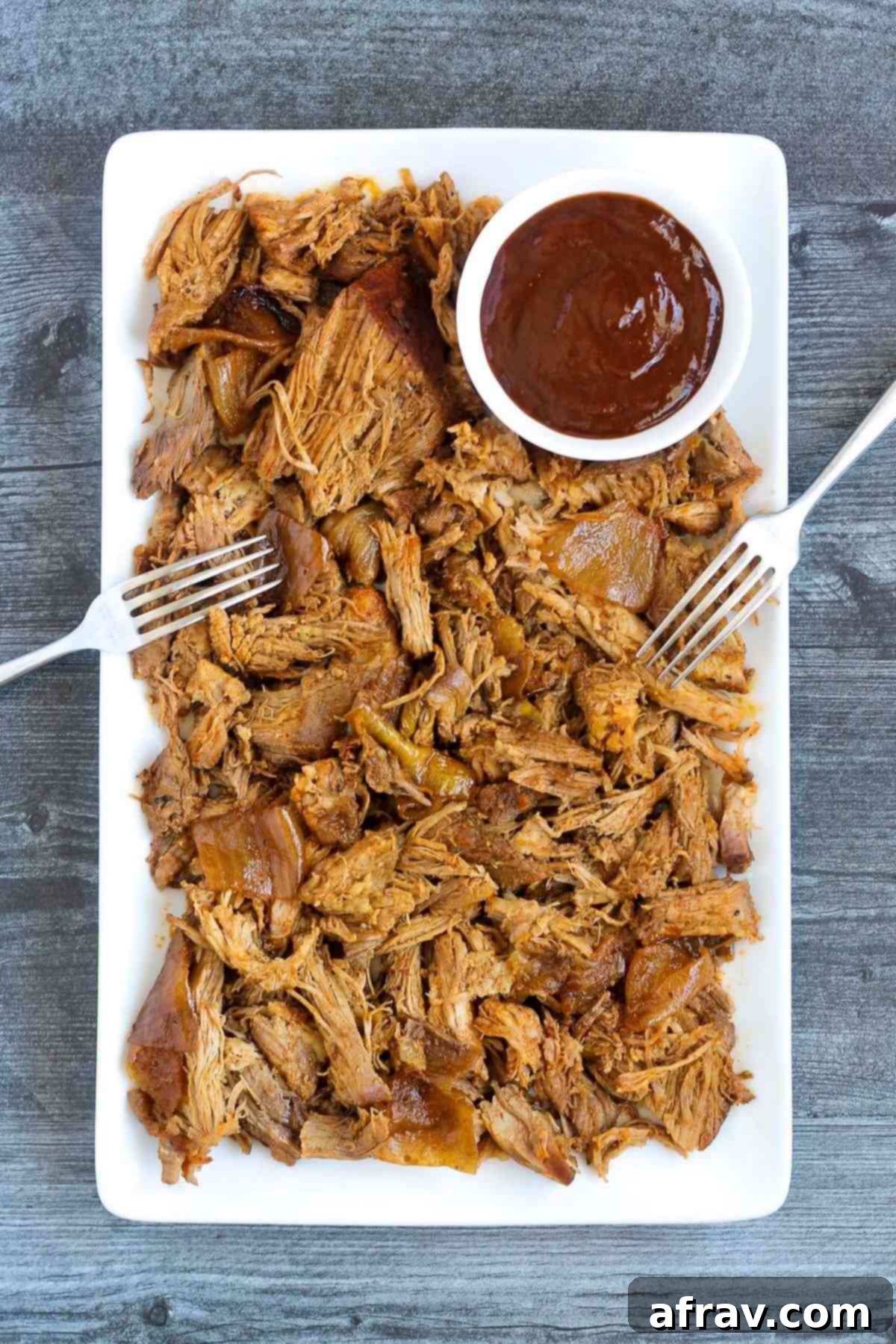 Pulled pork and a side of BBQ sauce with two forks on a serving tray.