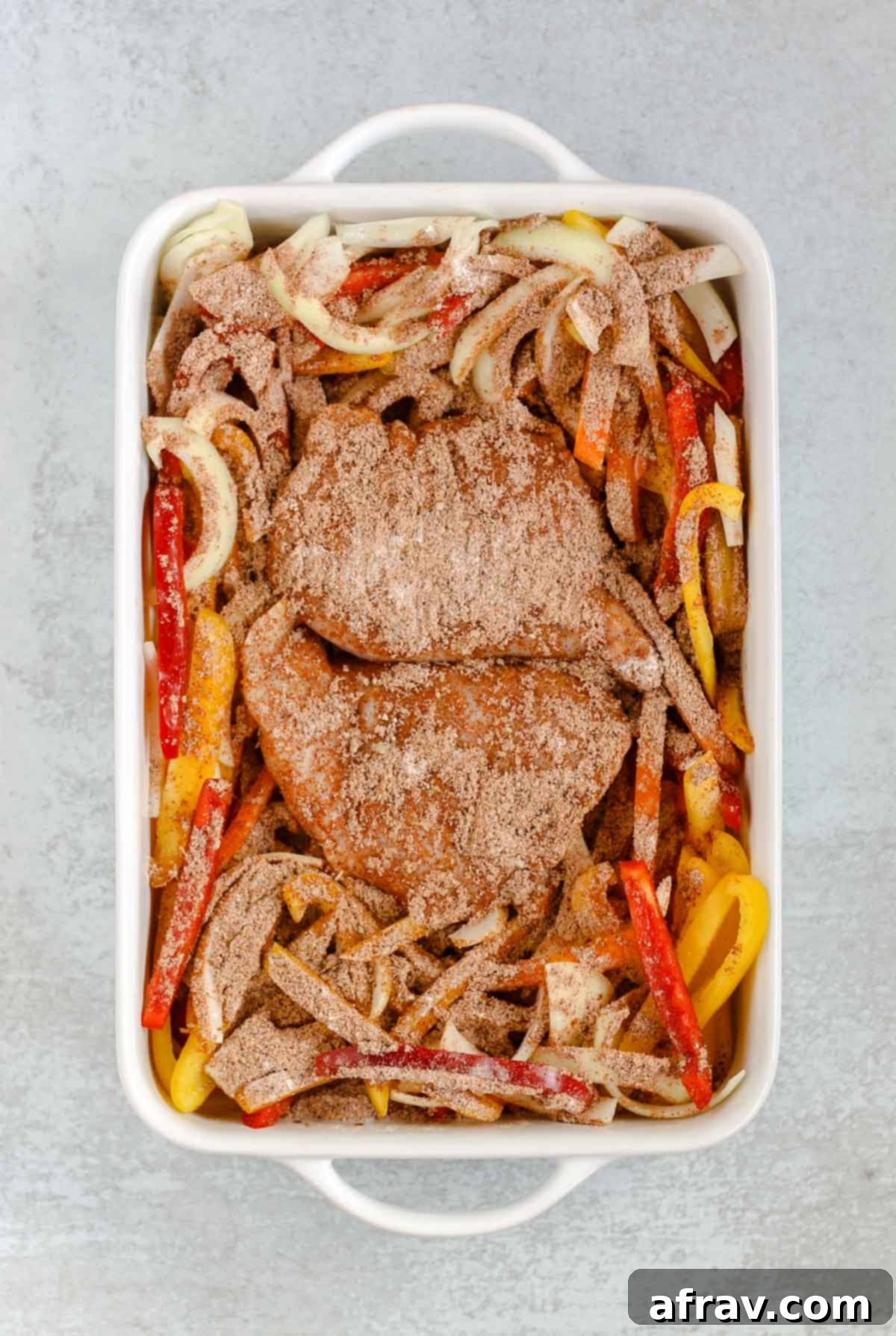 Sheet Pan Fajitas 4 Sliced bell peppers and onions arranged around chicken breasts in a white baking dish, ready for seasoning and baking.