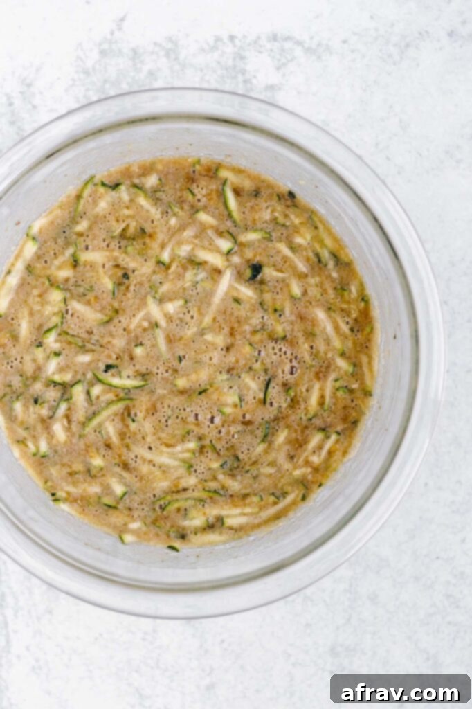 Grated zucchini and wet ingredients (banana, oil, sugar, eggs, vanilla) mixed in a glass bowl.