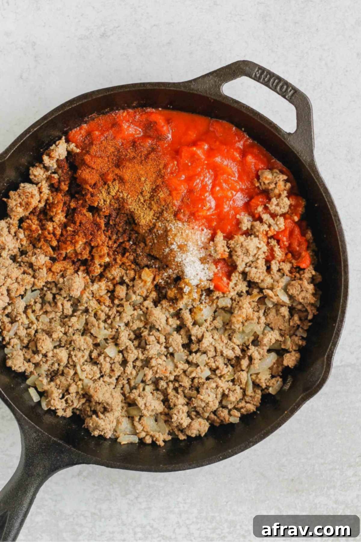Cooked ground beef, canned tomatoes, and seasoning in a cast iron skillet.