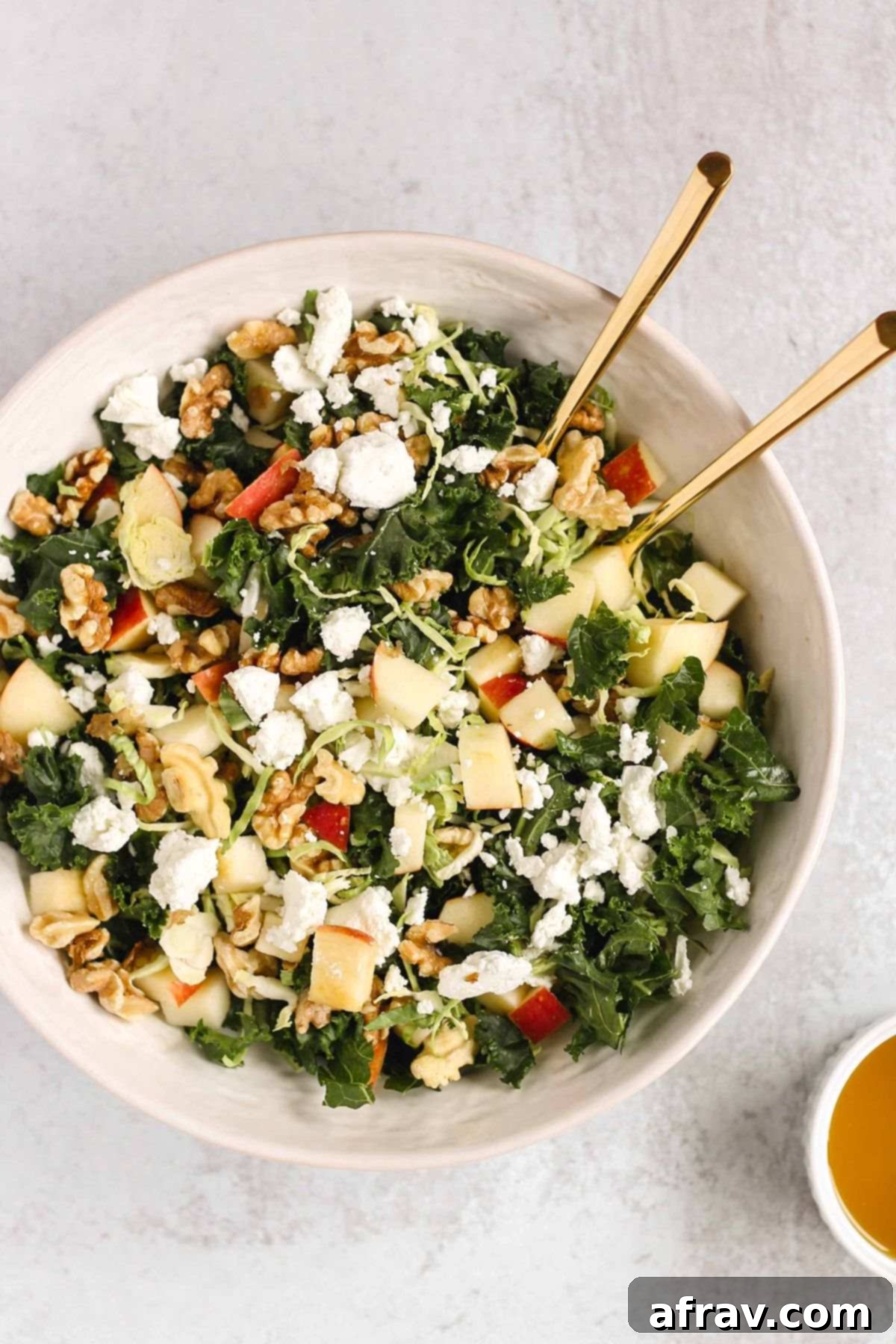 Kale and Brussels sprouts salad in a white serving bowl with gold salad utensils and a ramekin of dressing on the side.