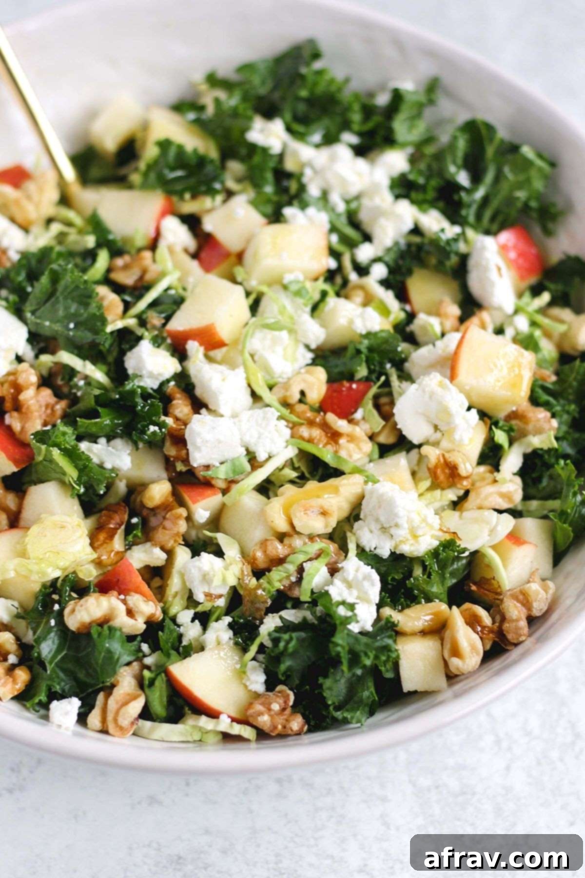 Kale salad with shaved Brussels sprouts topped with apples, walnuts, and goat cheese crumbles in a white serving bowl.