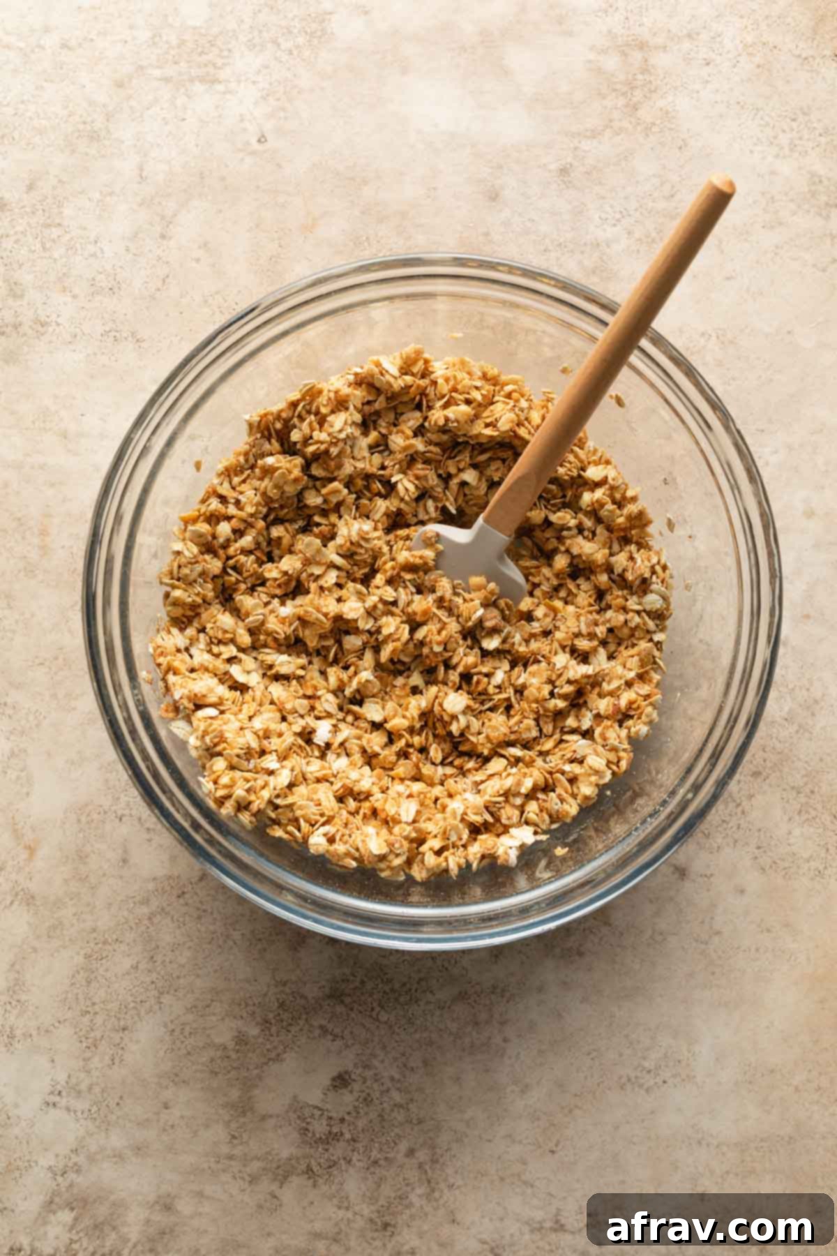 Granola mixture being stirred in a clear glass bowl with a rubber spatula, showing all ingredients combined.