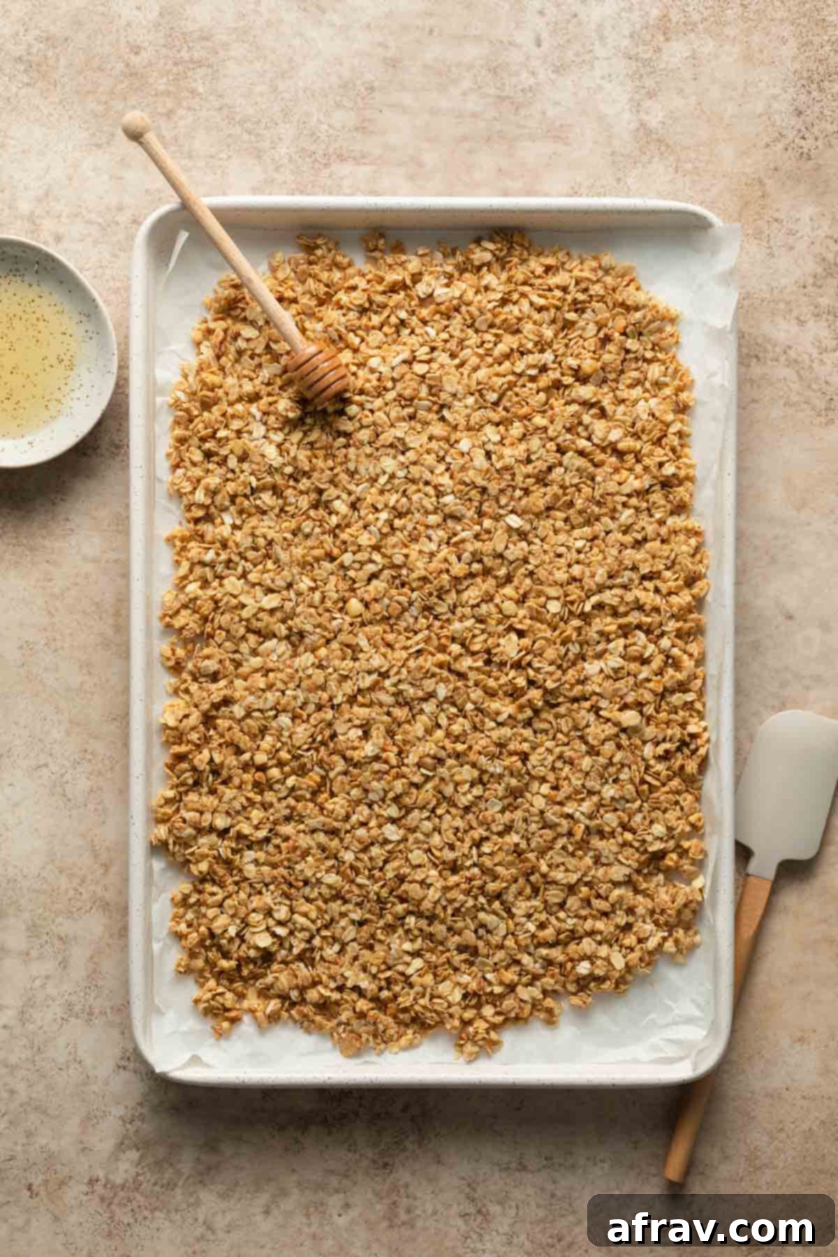 Granola mixture evenly spread and pressed onto a white baking sheet lined with parchment paper, ready for baking.