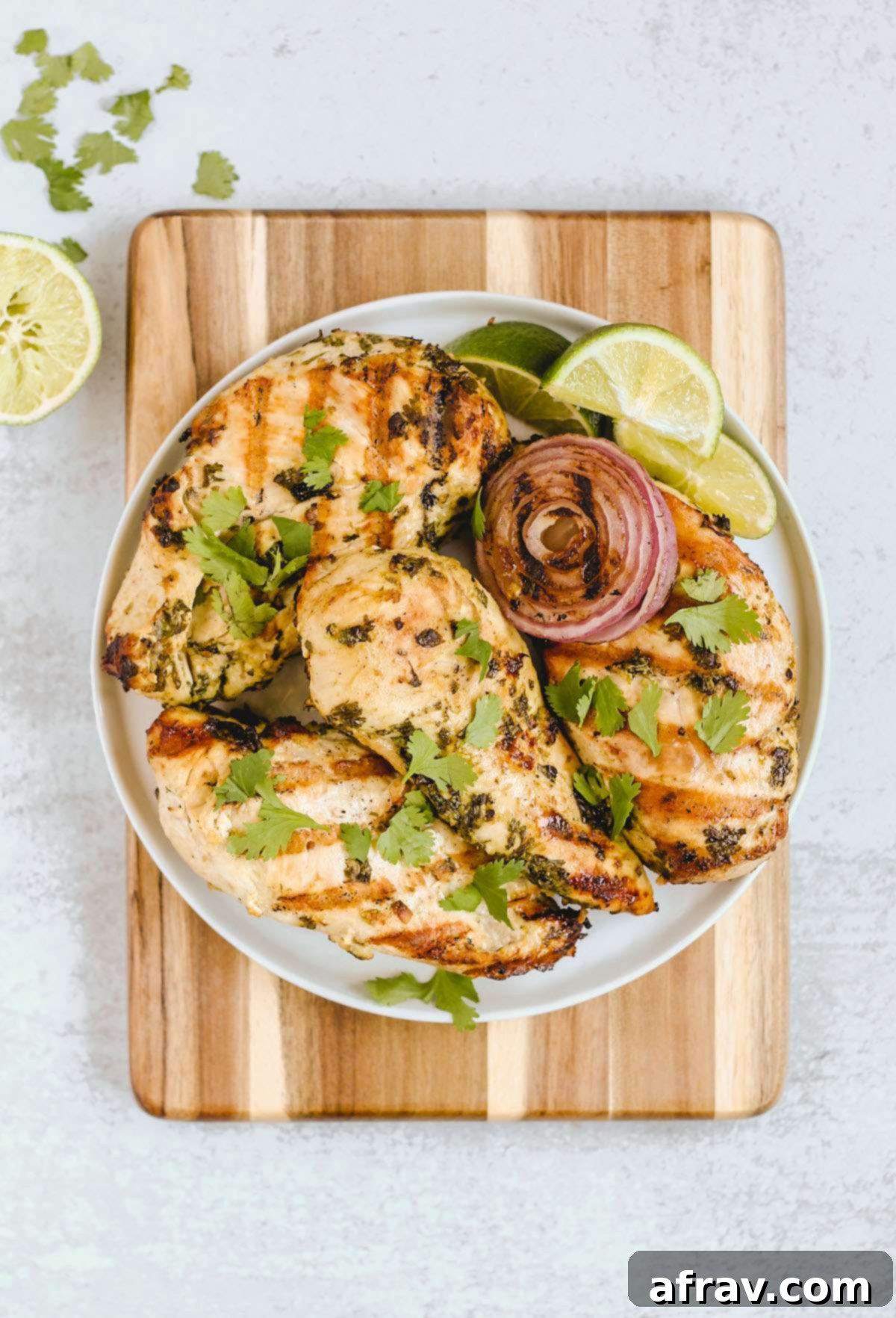 Four grilled cilantro lime chicken breasts with grilled onions and lime slices on a round plate on top of a wood cutting board.