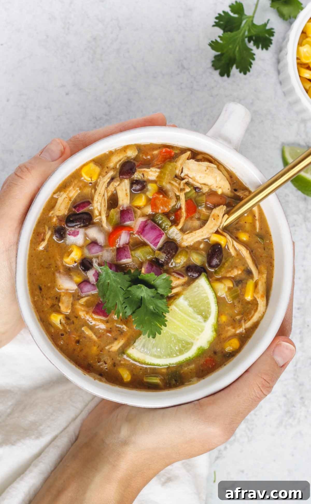 Hands holding a bowl of chicken chili topped with fresh cilantro and a lime wedge, highlighting its comforting and fresh appeal.