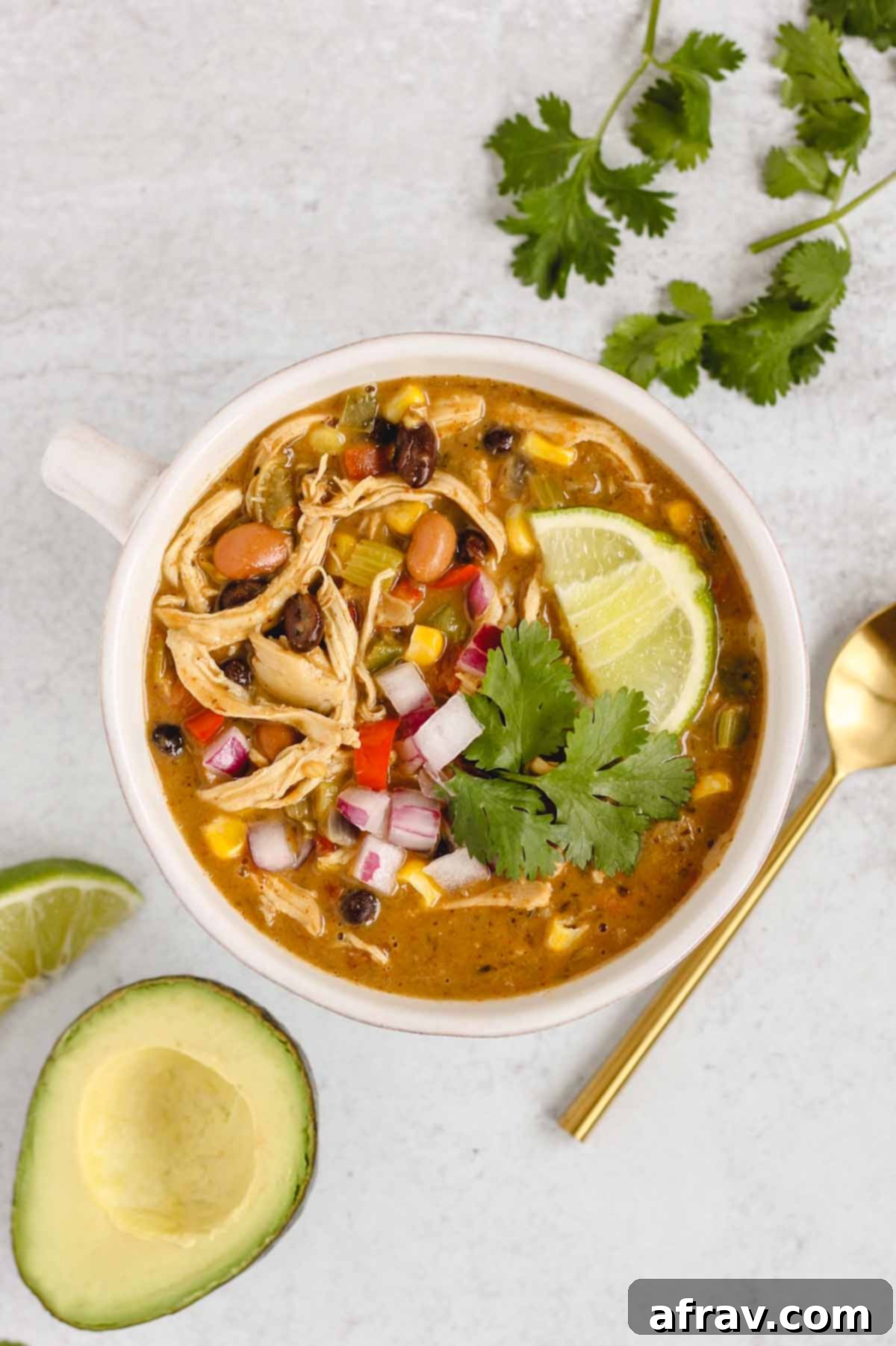 A beautifully presented bowl of Southwest Chicken Chili, garnished with fresh cilantro and a lime wedge, accompanied by a gold spoon and an avocado half on the side.