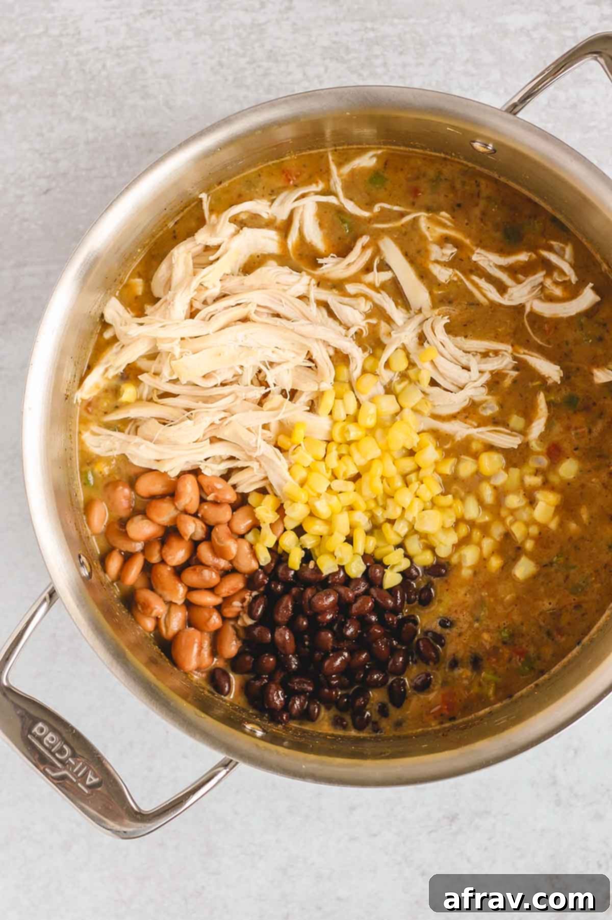 Shredded chicken, black beans, pinto beans, and corn added to the rich chili base in a stainless steel pot, creating a hearty mixture.