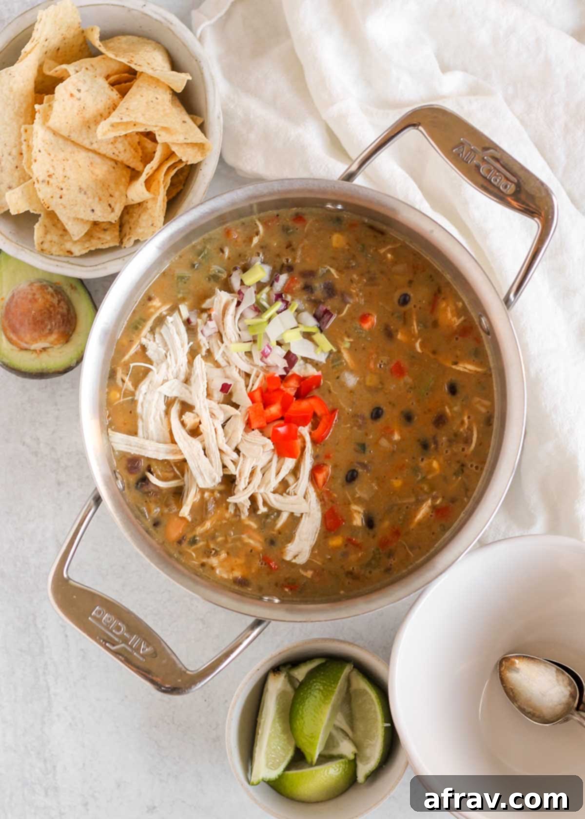 A large stainless steel stock pot filled with rich Southwest Chicken Chili, surrounded by bowls, lime wedges, fresh avocado, and tortilla chips, ready for a feast.