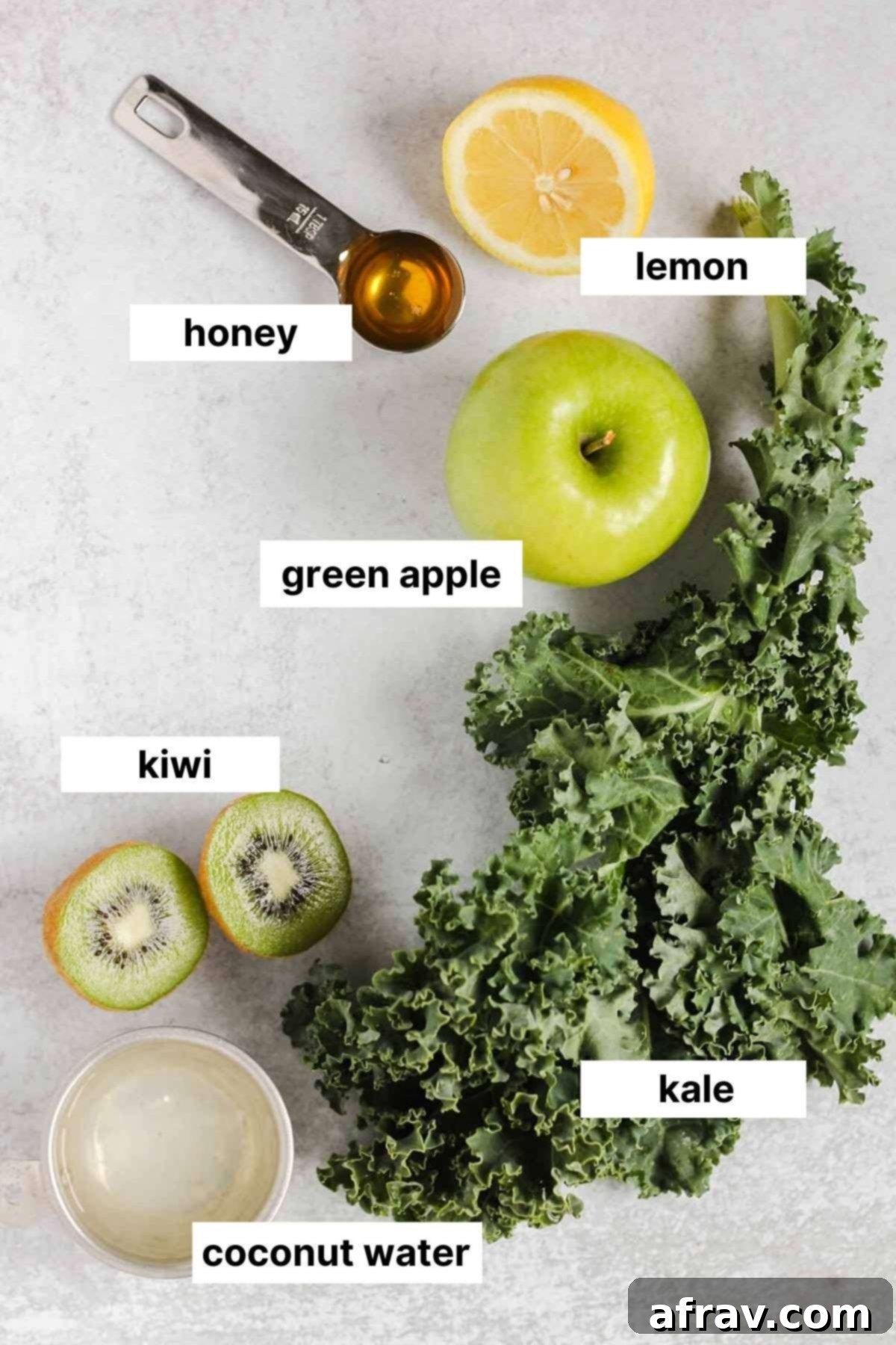 A flat lay photograph showcasing the simple, fresh ingredients required for the kale smoothie: fresh kale leaves, a green apple, a kiwi, a lemon, a small jar of honey, and a bottle of coconut water.