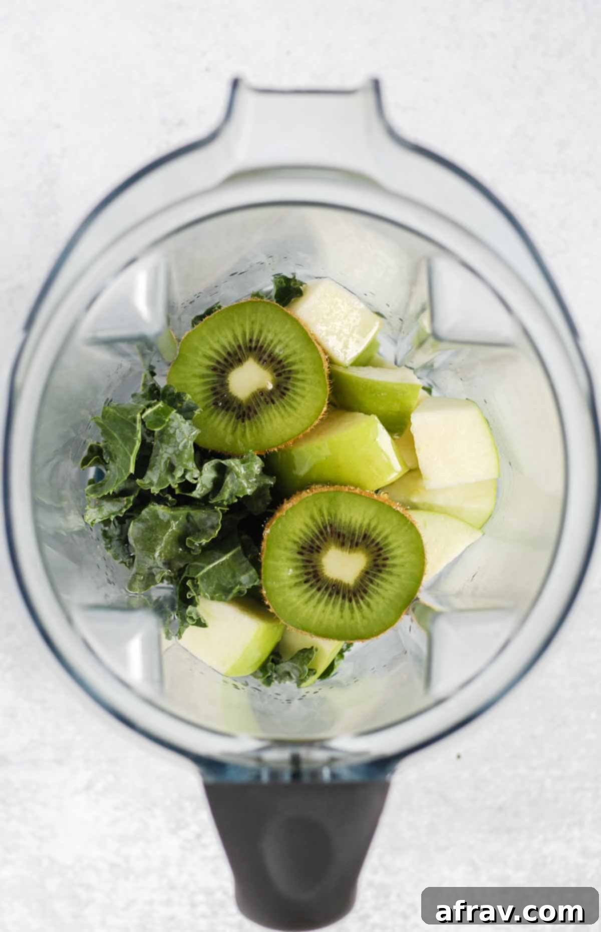Fresh kale, apple, and kiwi, along with lemon and honey, are placed inside a blender jar, ready to be processed into a smoothie.