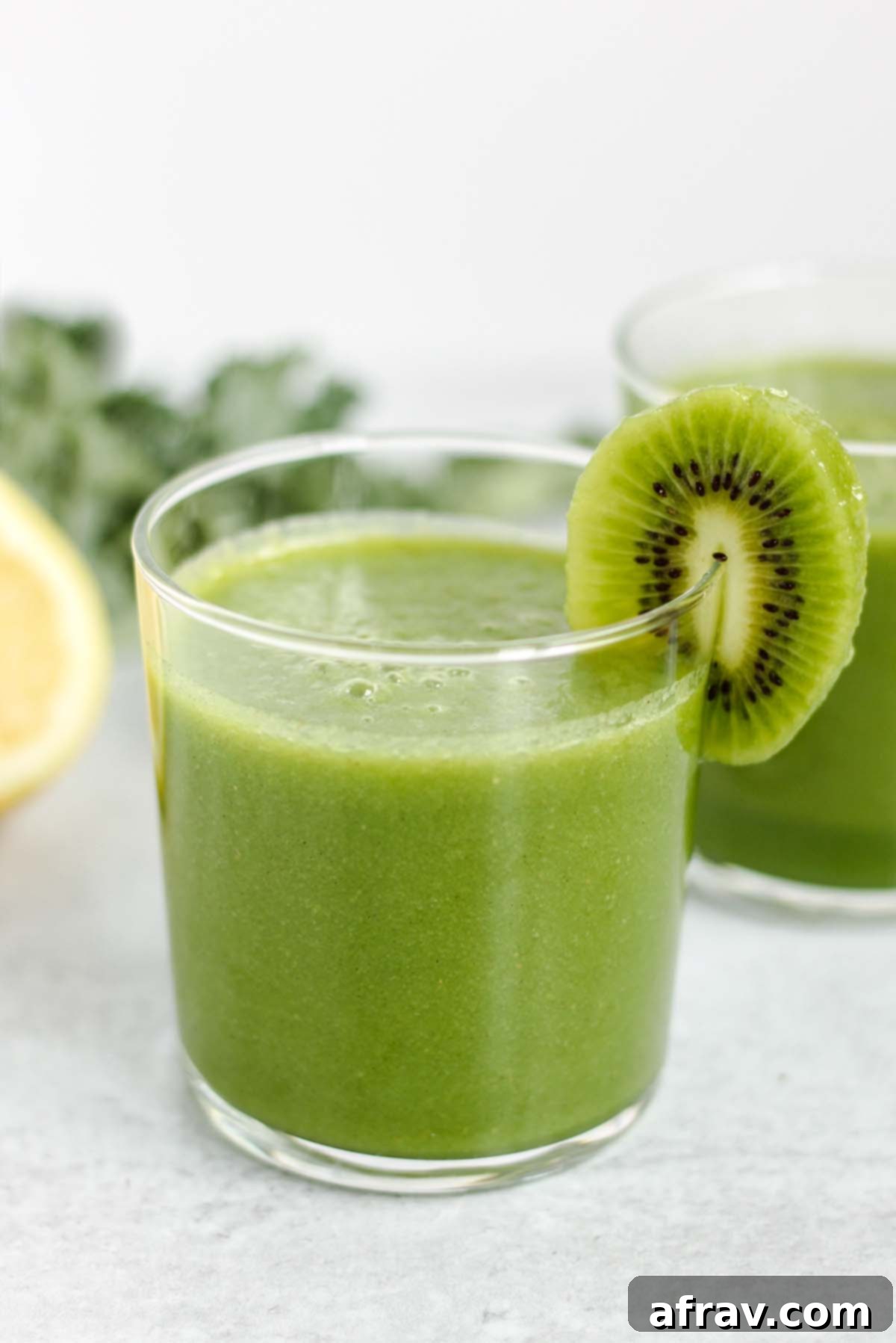 A small, elegant glass filled with a vibrant green kale apple smoothie, garnished with a fresh slice of kiwi on the rim of the glass.