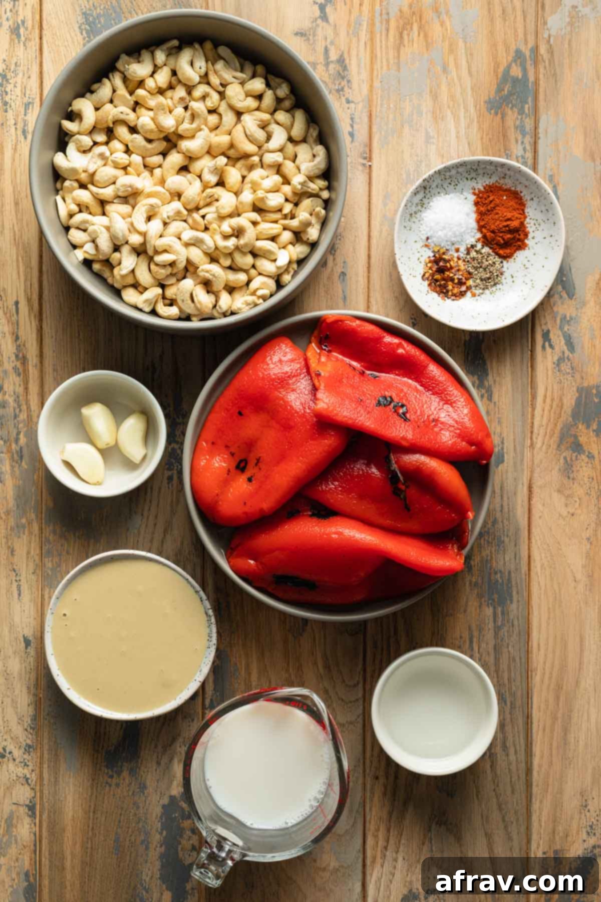 A selection of fresh ingredients laid out on a wooden surface, including jarred roasted red peppers, raw cashews, a bottle of non-dairy milk, a jar of tahini, garlic cloves, and various spices, ready for making the roasted red pepper dip.