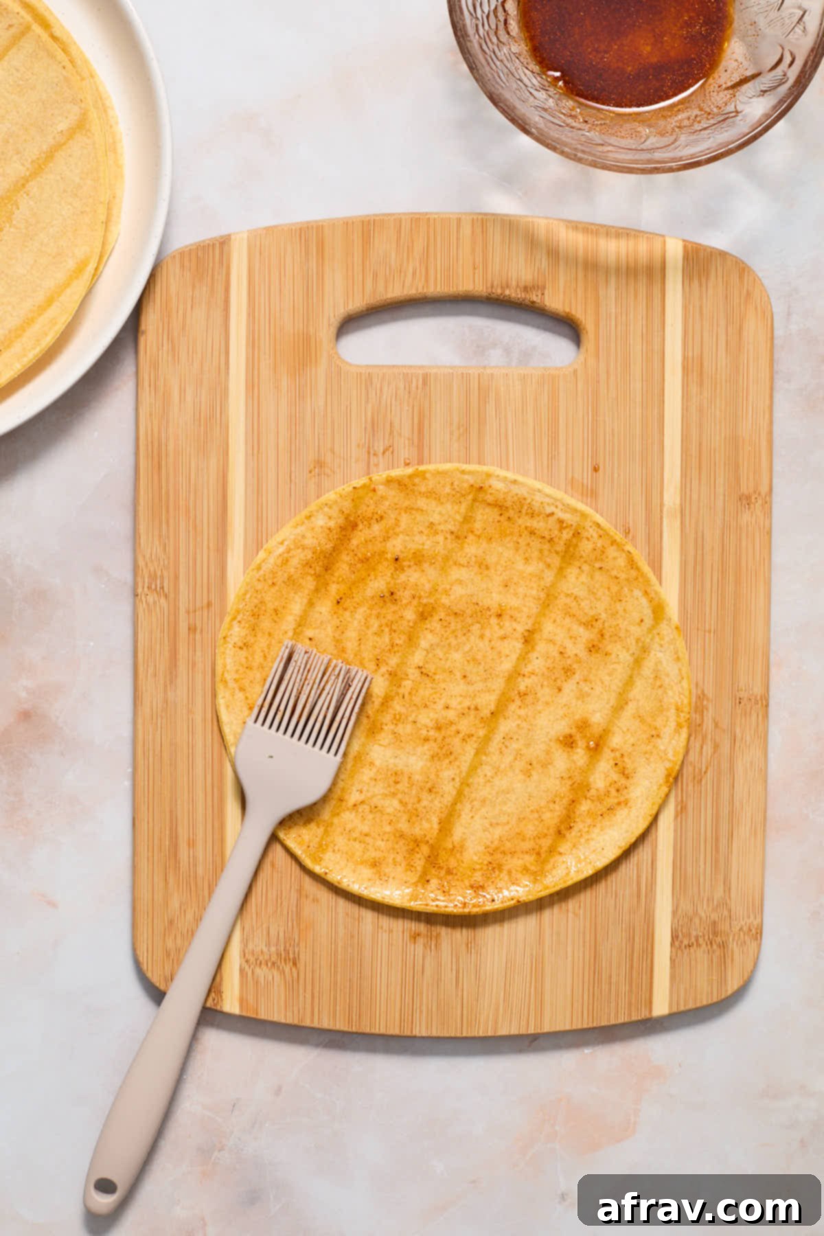 A corn tortilla on a wooden board being brushed with the oil mixture.