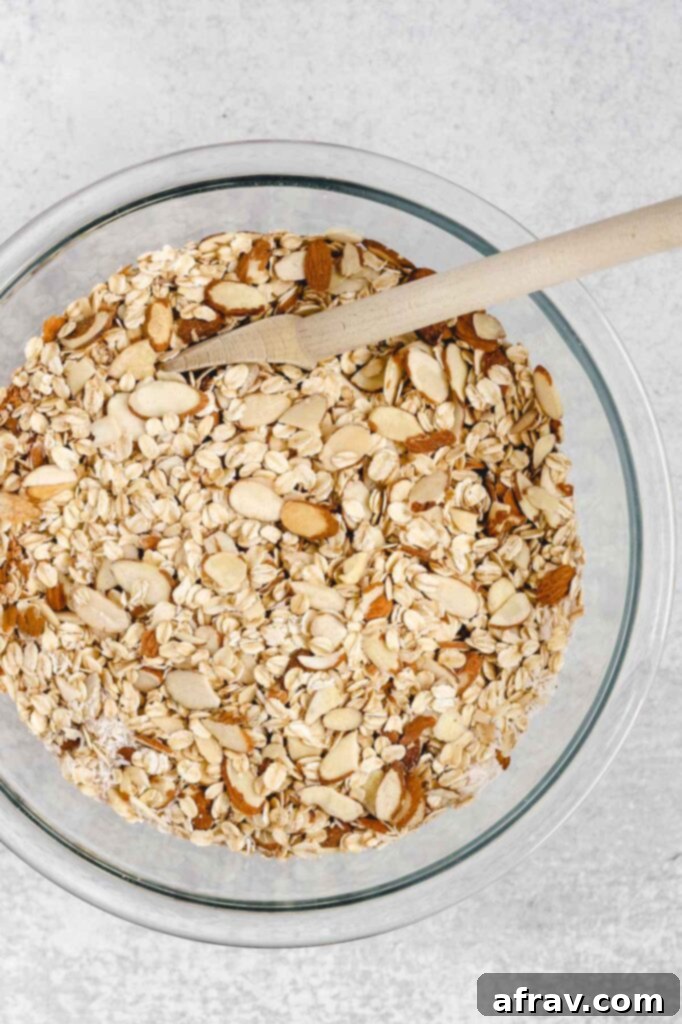 Dry ingredients for oat and honey granola, including oats, almonds, coconut, and cinnamon, in a clear glass mixing bowl.