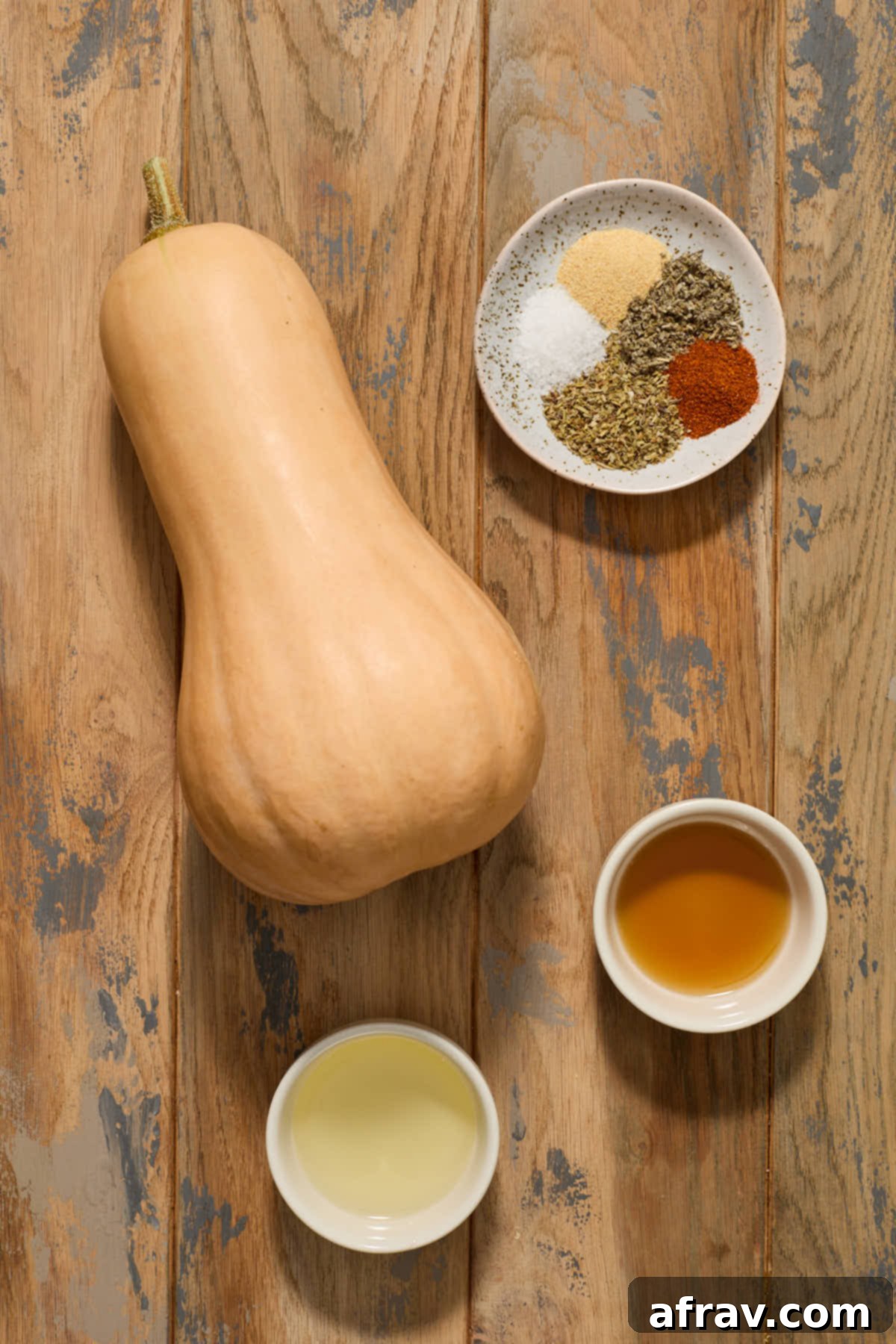 Individual ingredients for air fryer butternut squash, including a whole squash, olive oil, maple syrup, and various seasonings, neatly arranged on a wooden surface.