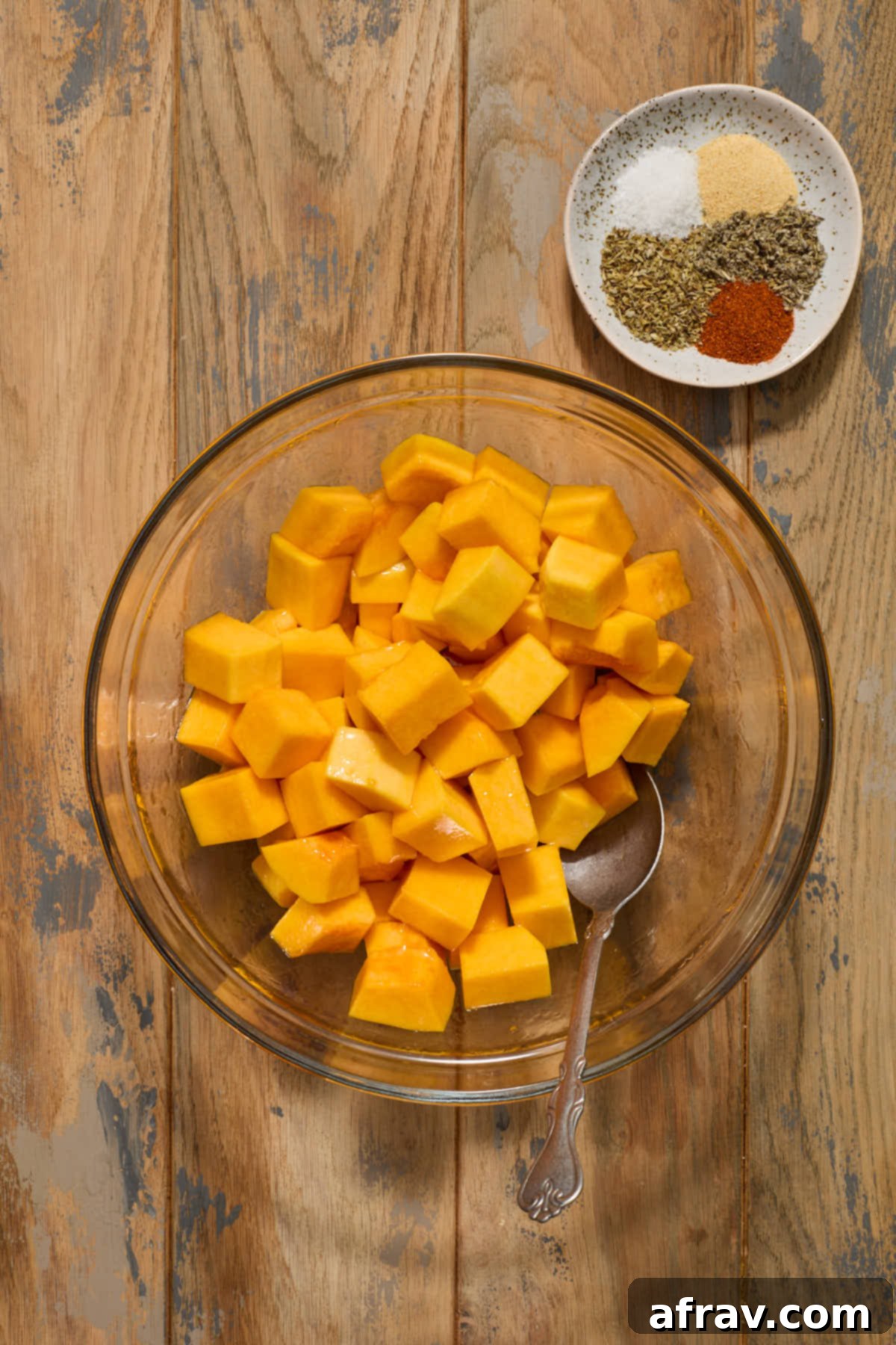Butternut squash cubes being tossed in a bowl with olive oil and maple syrup, with seasonings waiting on the side.