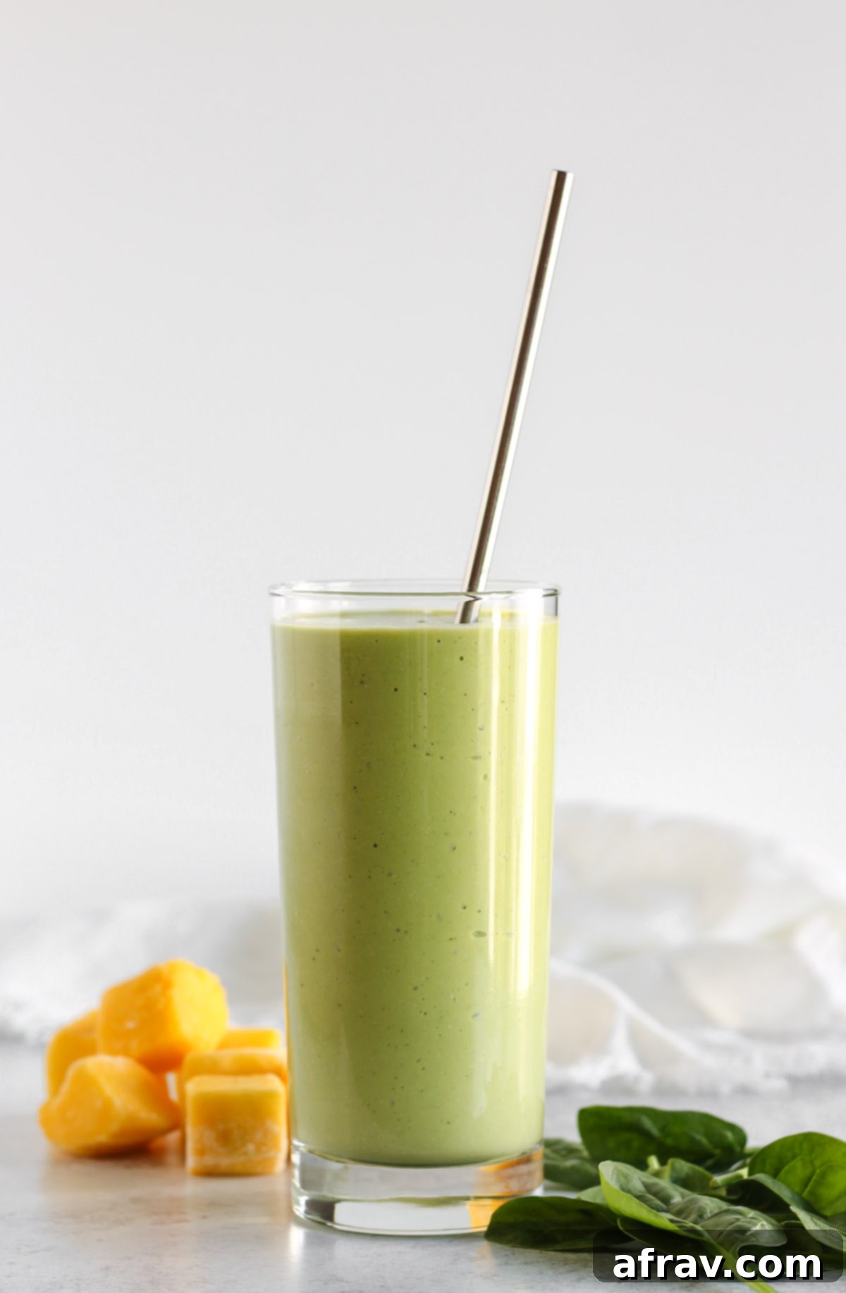 Spinach mango smoothie in a tall glass with a stainless steel straw, showcasing its vibrant green color and creamy texture.
