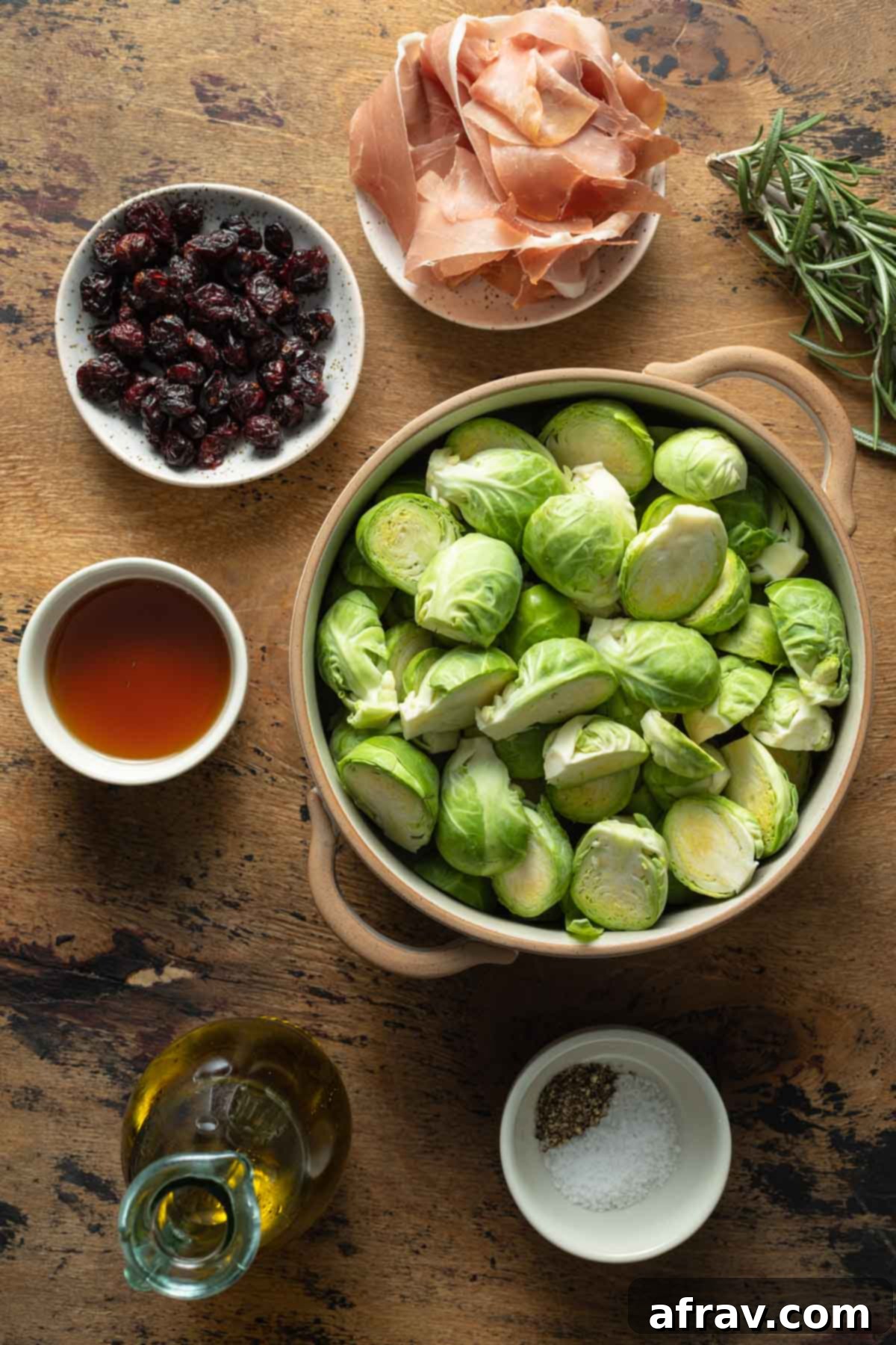 Ingredients for roasted Brussels sprouts arranged in individual bowls, showing fresh rosemary, maple syrup, cranberries, oil, and prosciutto.