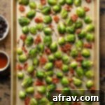Roasted Brussels sprouts with prosciutto on a baking sheet with dried cranberries and maple syrup off to the side, ready to be tossed.