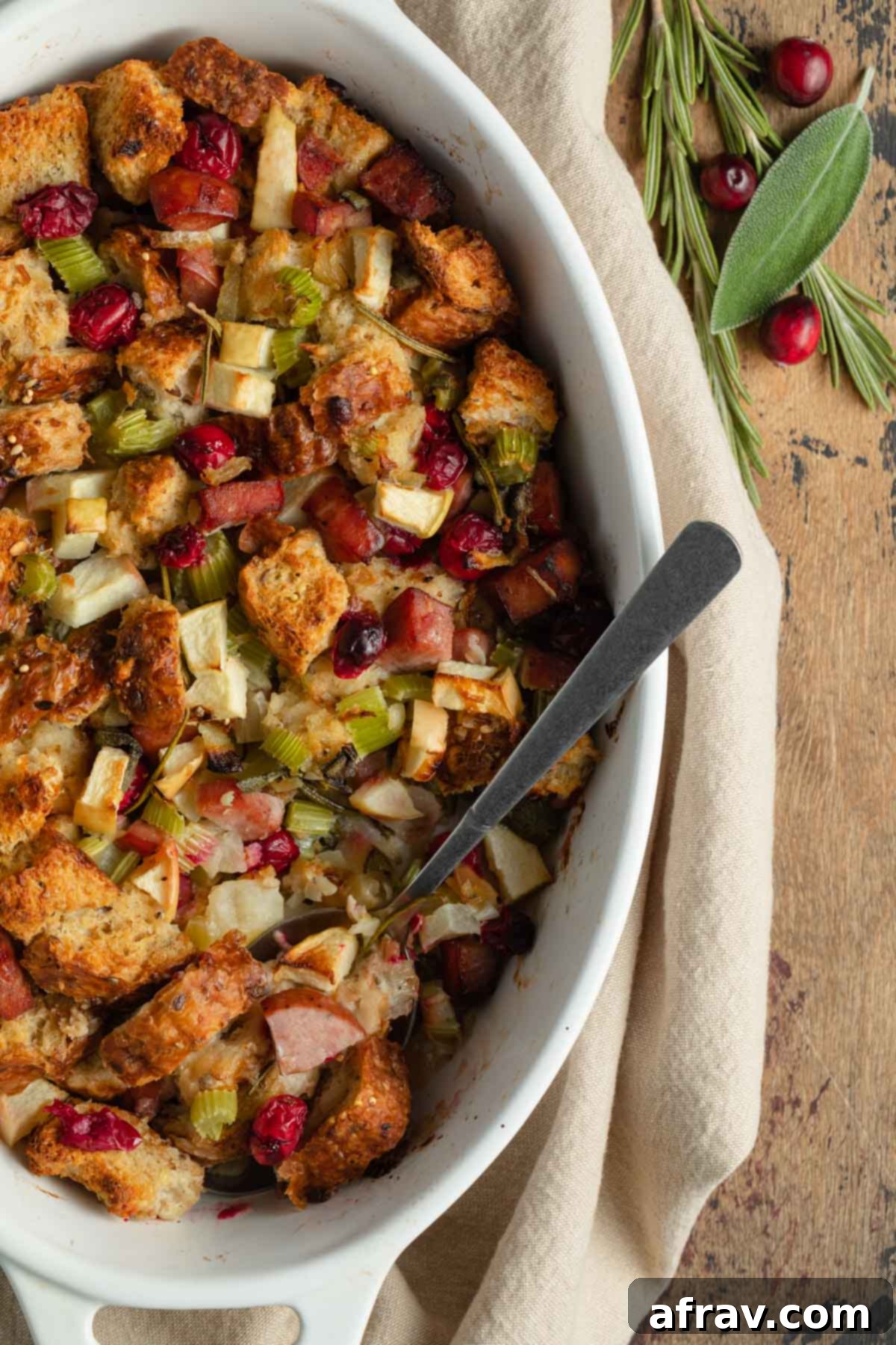 Close up view of whole wheat stuffing in a white dish with a serving spoon inserted into it, highlighting the crispy edges and festive cranberries.