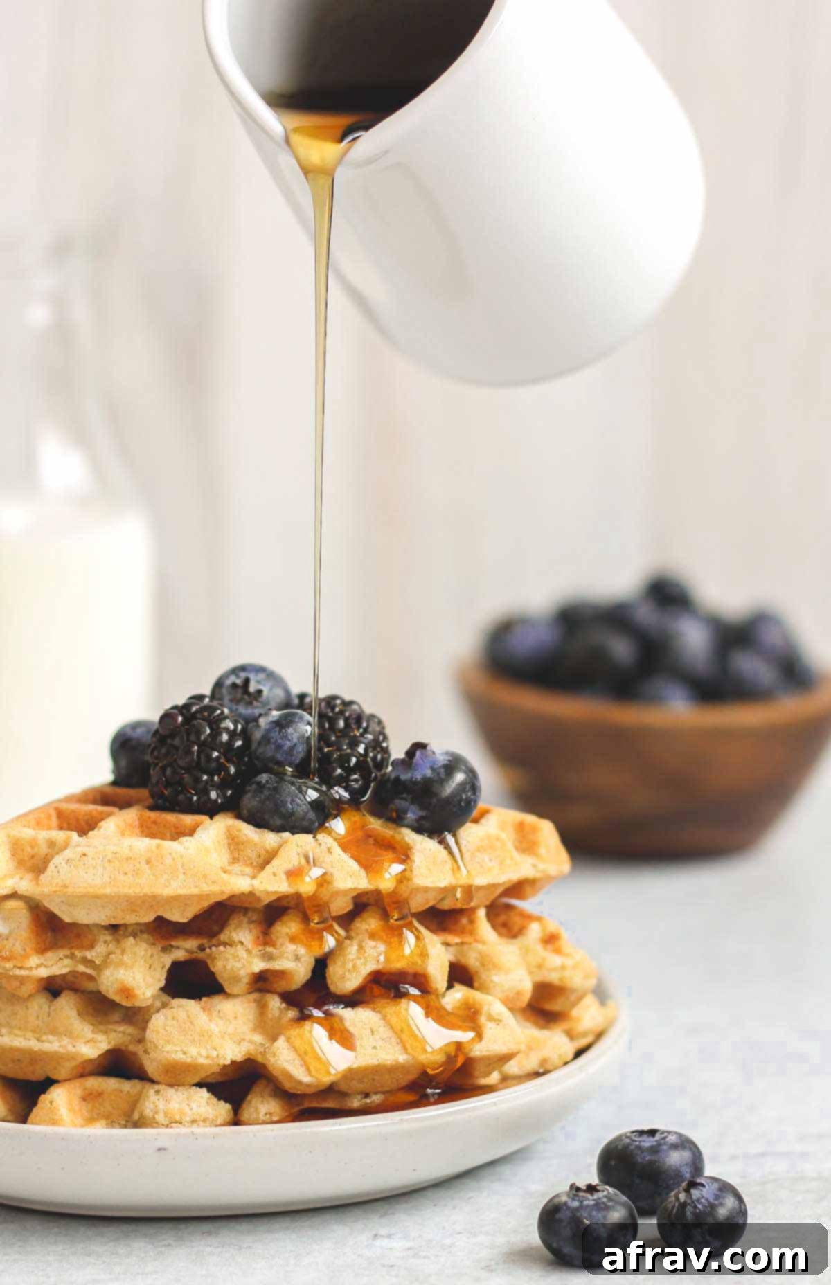 Syrup being poured over a stack of waffles topped with fresh blackberries and blueberries.