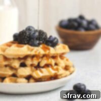 Syrup being poured over a stack of waffles topped with fresh blackberries and blueberries.