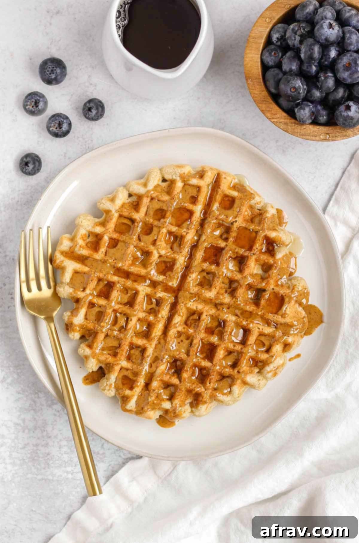 Deliciously Free Waffles 8 A single gluten-free waffle on a white plate, beautifully drizzled with almond butter and syrup, adorned with fresh blueberries and a small bowl of additional syrup.