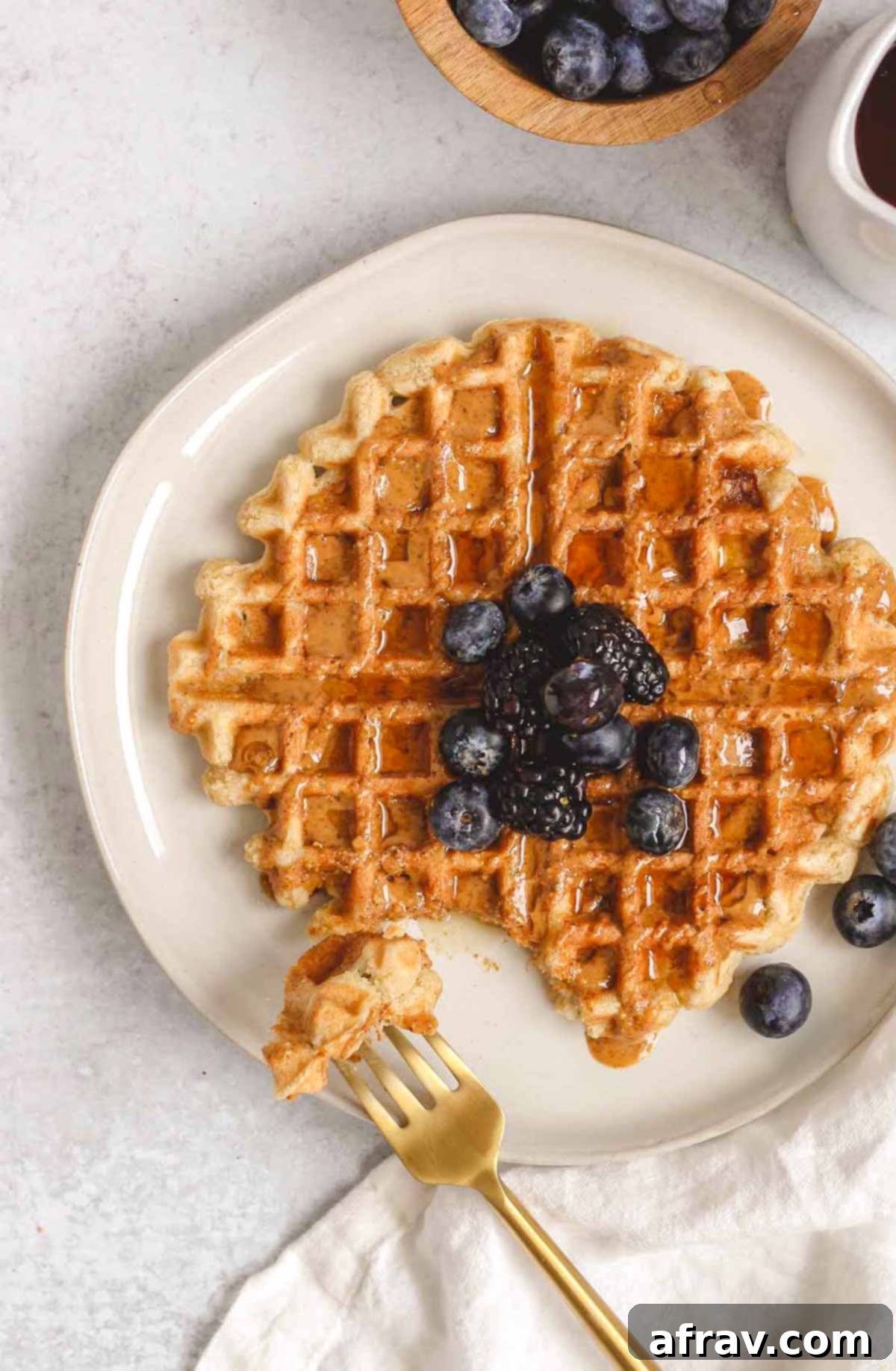 Deliciously Free Waffles 9 A single gluten-free waffle topped with an assortment of berries, drizzled with almond butter and syrup, with a gold fork taking a bite.