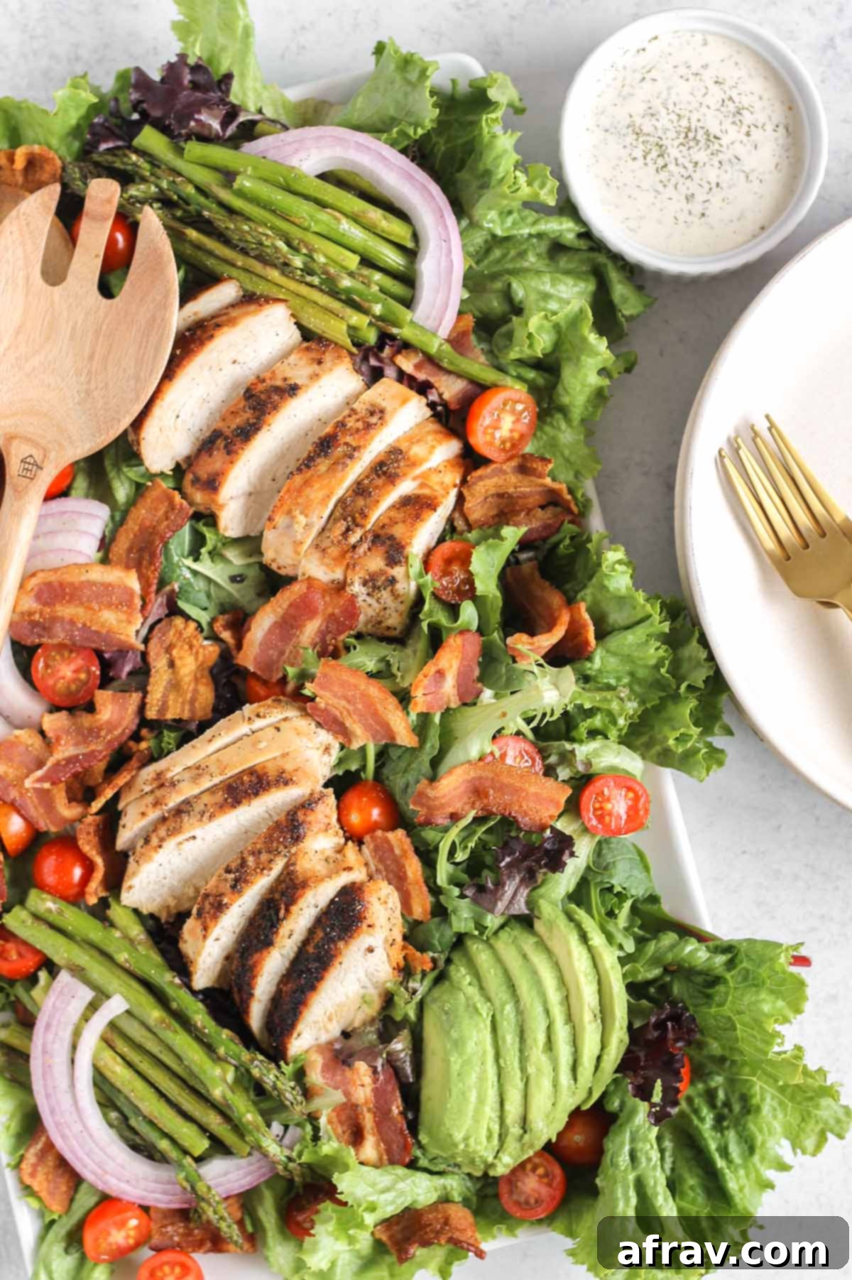 BLT Bliss Chicken Salad 4 A large, rustic wooden serving tray abundantly filled with a vibrant BLT chicken salad, featuring glistening chicken strips, crunchy bacon bits, bright red tomatoes, green asparagus spears, and creamy avocado, with a small dish of ranch dressing on the side.