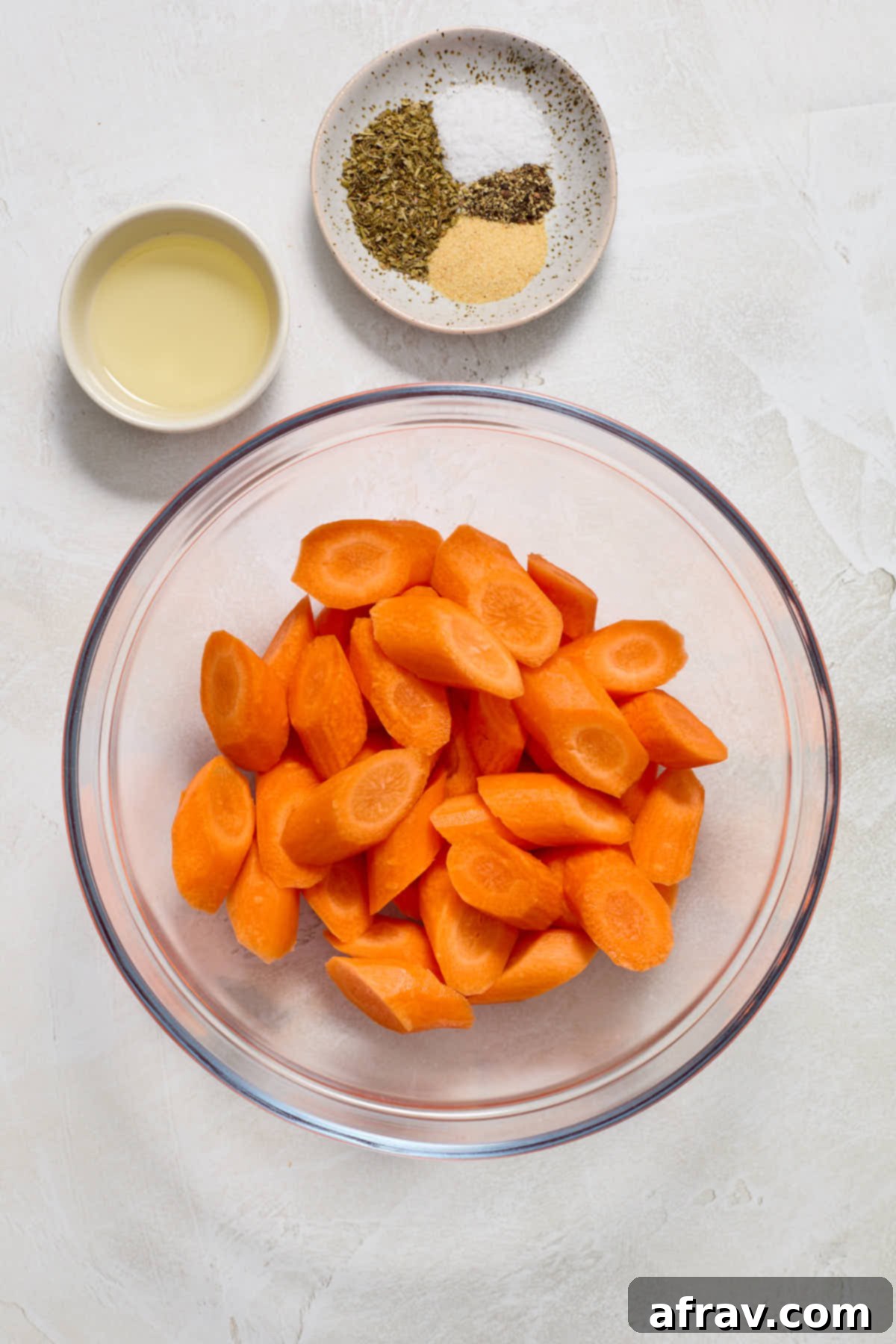 Peeled, washed, and sliced carrots in a glass bowl, with olive oil and various seasonings ready on the side.