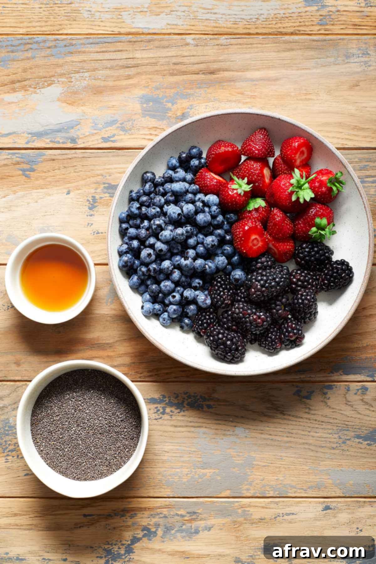 Individual bowls of fresh strawberries, blueberries, blackberries, chia seeds, and maple syrup arranged on a wooden table, showcasing the ingredients for chia jam.