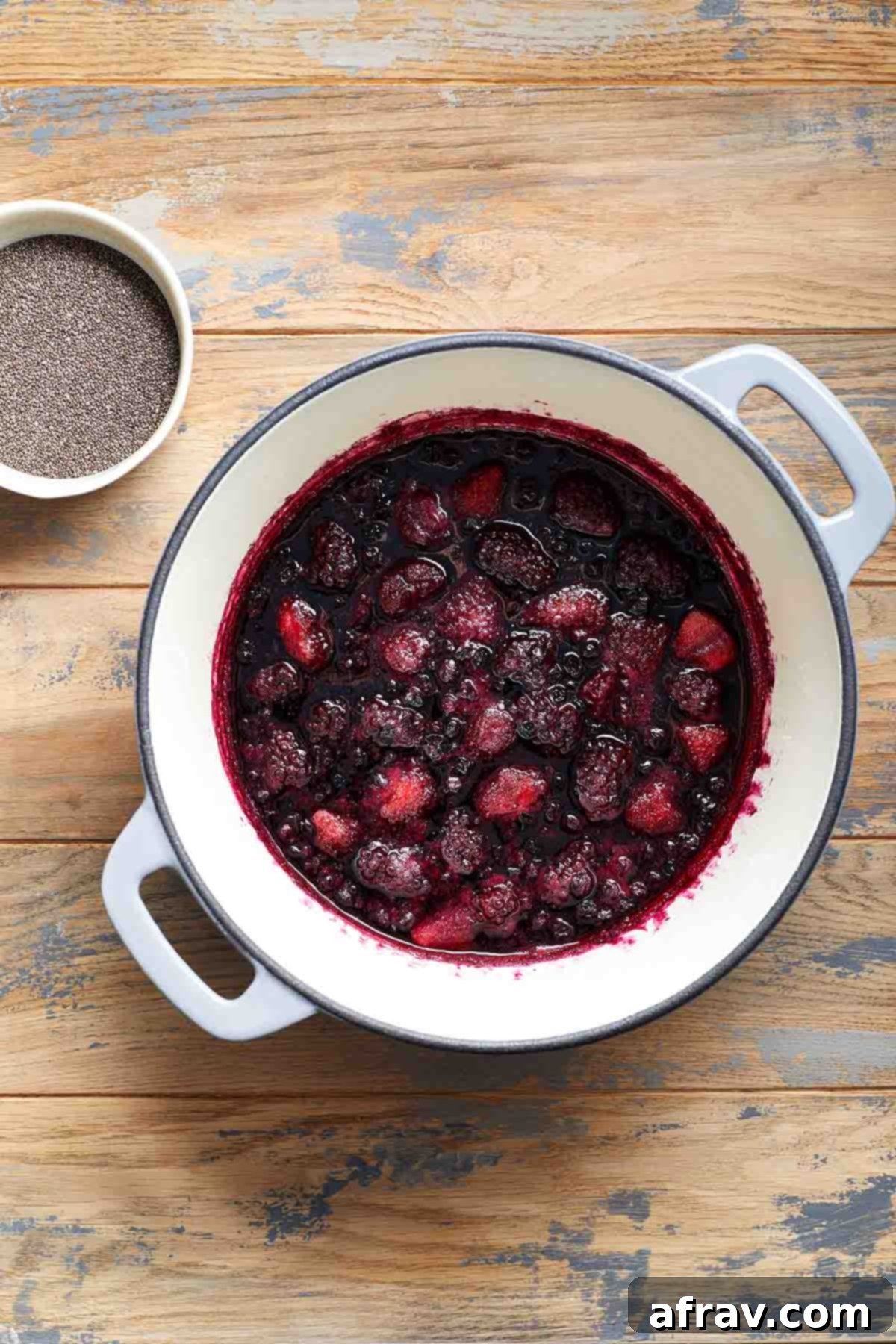 Berries cooking down in a pot with maple syrup, next to a bowl of chia seeds.