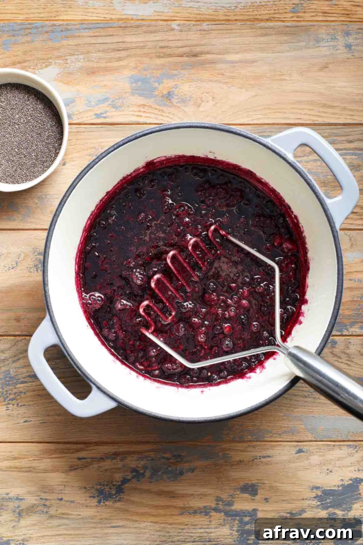 Mashed berries in a pot with a potato masher inside, showing the texture of the cooked fruit.