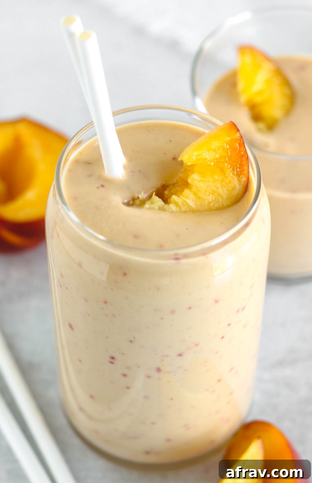 Tall glass filled with a peach banana smoothie topped with a peach slice and two straws.
