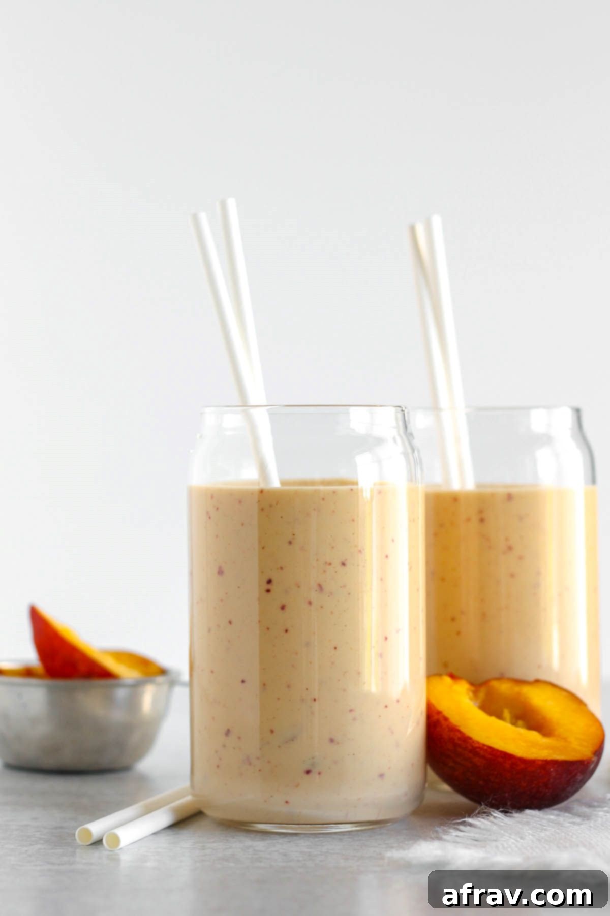 Two glasses filled with peach banana smoothie with two straws in each glass and half a peach laying next to the glasses.