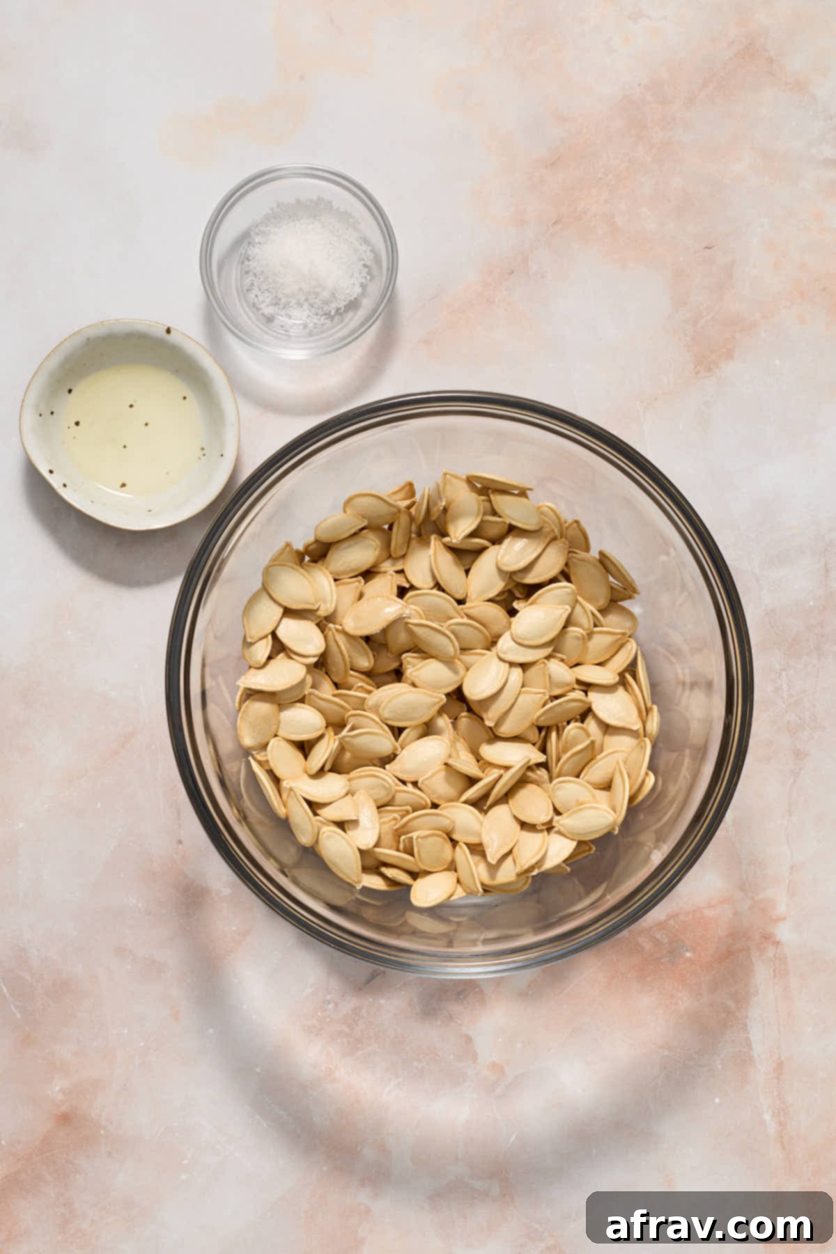 Fully dried pumpkin seeds in a bowl, with a small dish of olive oil and a small dish of sea salt next to it.