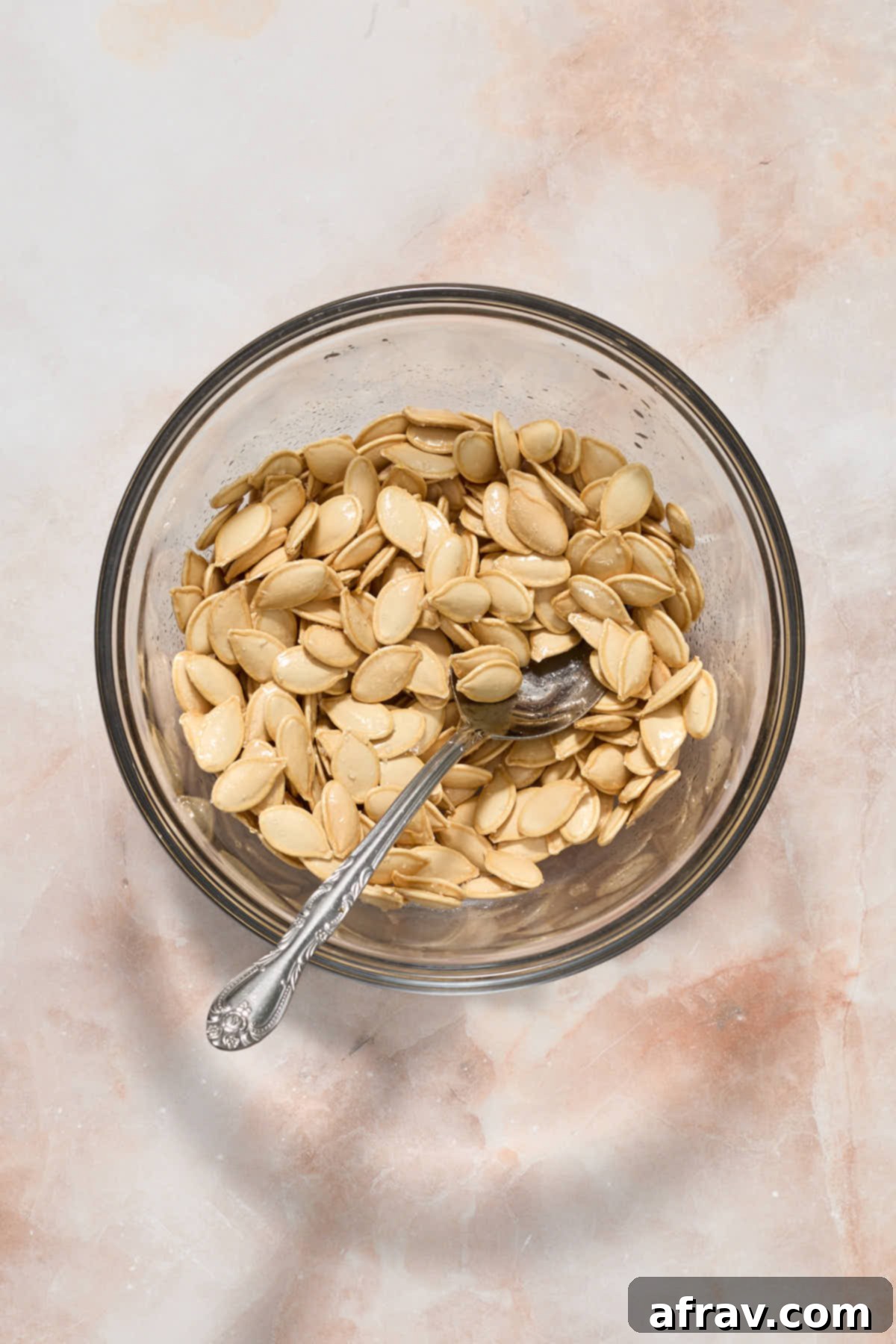 Seasoned pumpkin seeds glistening with oil and salt in a glass bowl, stirred with a spoon.