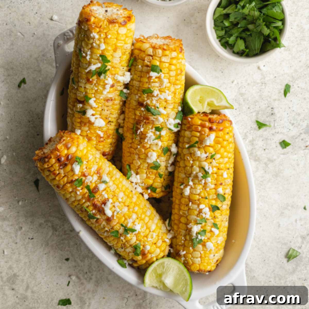 Effortless Air Fryer Side Dishes 15 Four ears of air fried corn in a white dish and topped with feta and parsley, resembling grilled corn.