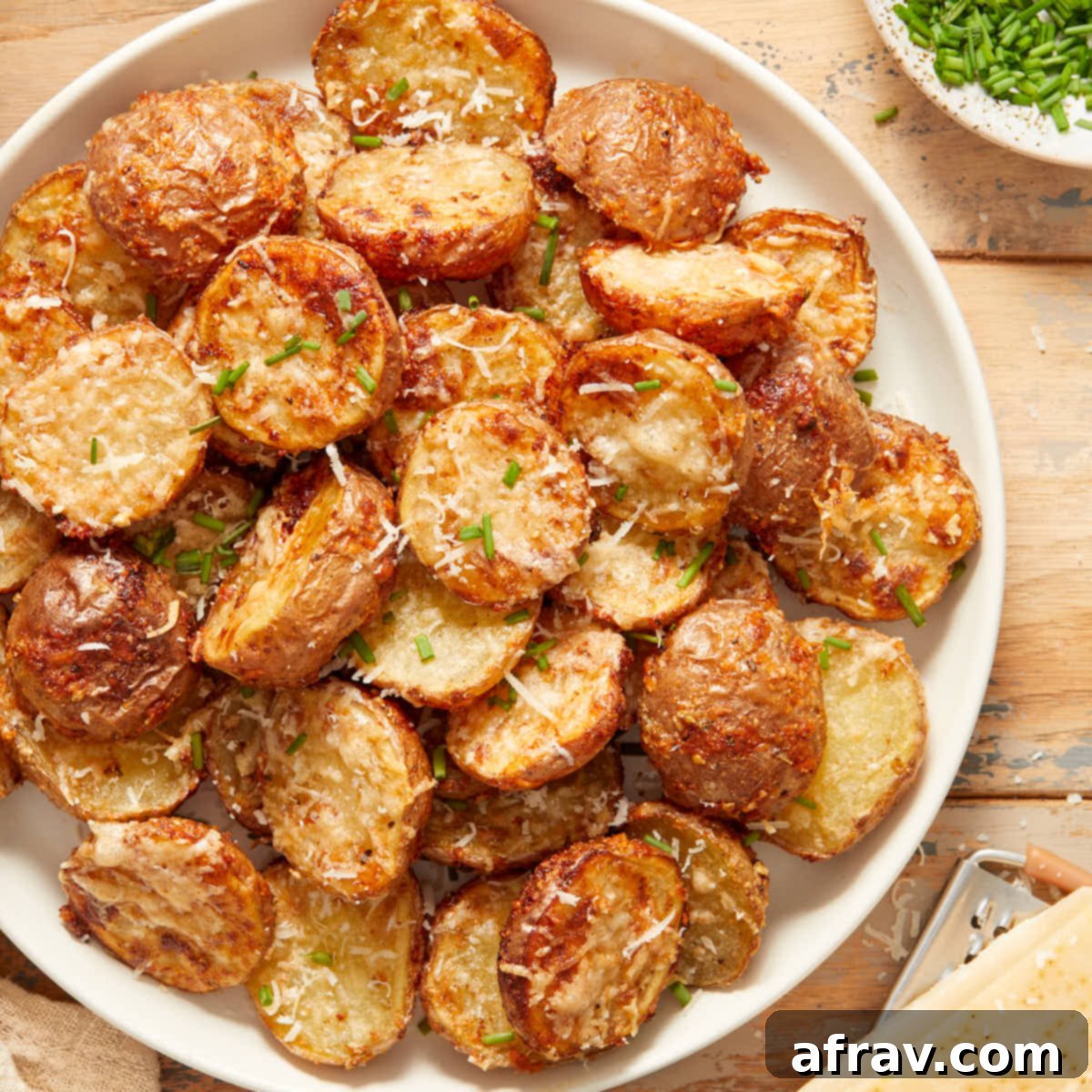 Effortless Air Fryer Side Dishes 5 Overhead view of air fryer parmesan potatoes on a white plate next to chopped chives and parmesan cheese, highlighting their crispy crust.