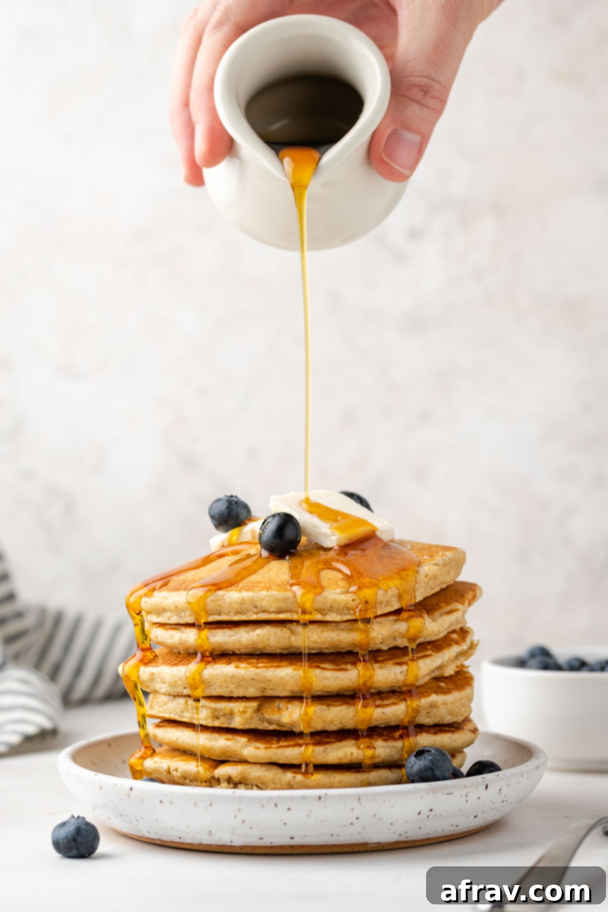 Syrup being poured over a stack of oat flour pancakes topped with blueberries.
