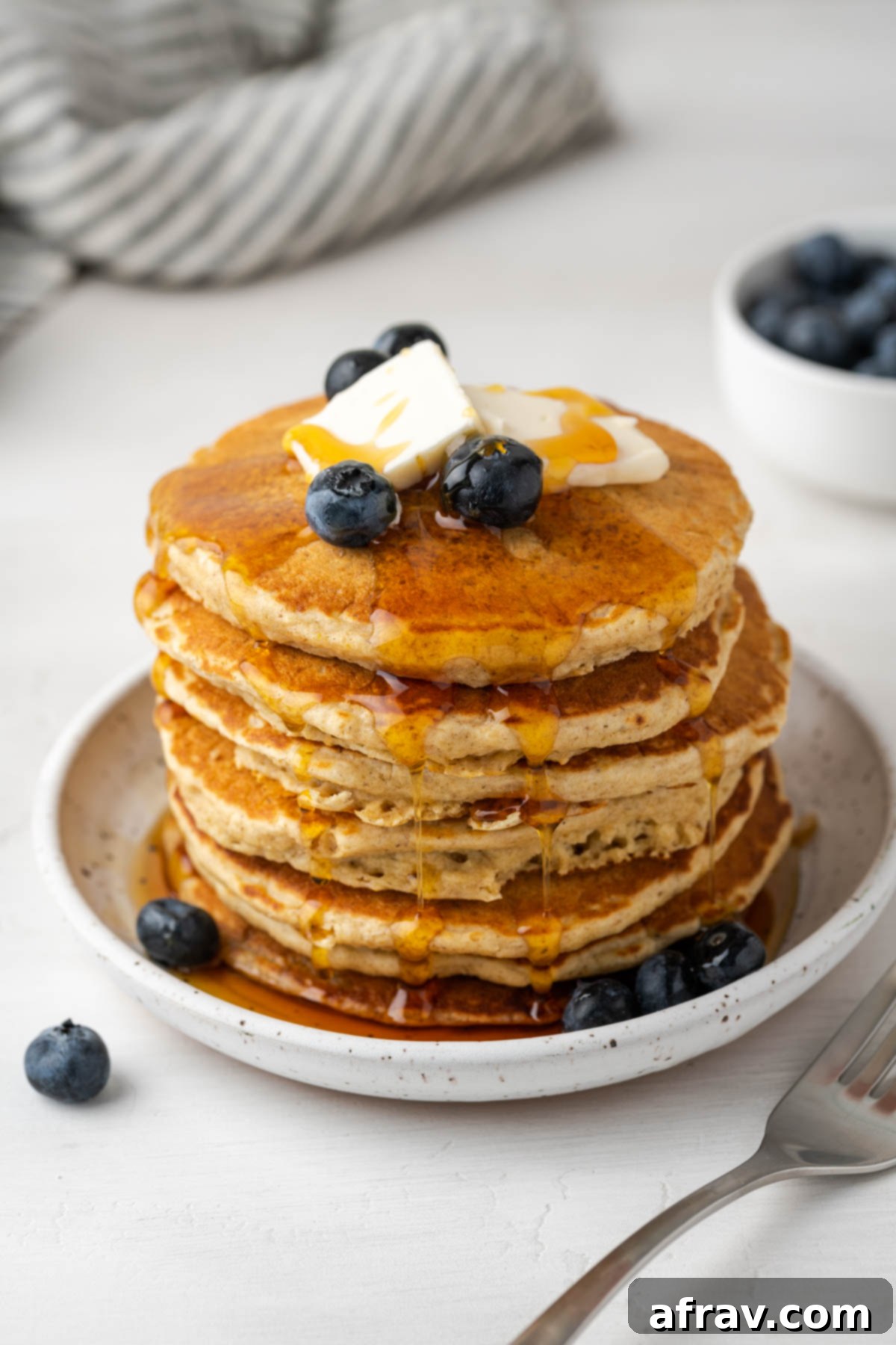Stack of pancakes topped with butter squares, blueberries, and dripping syrup.