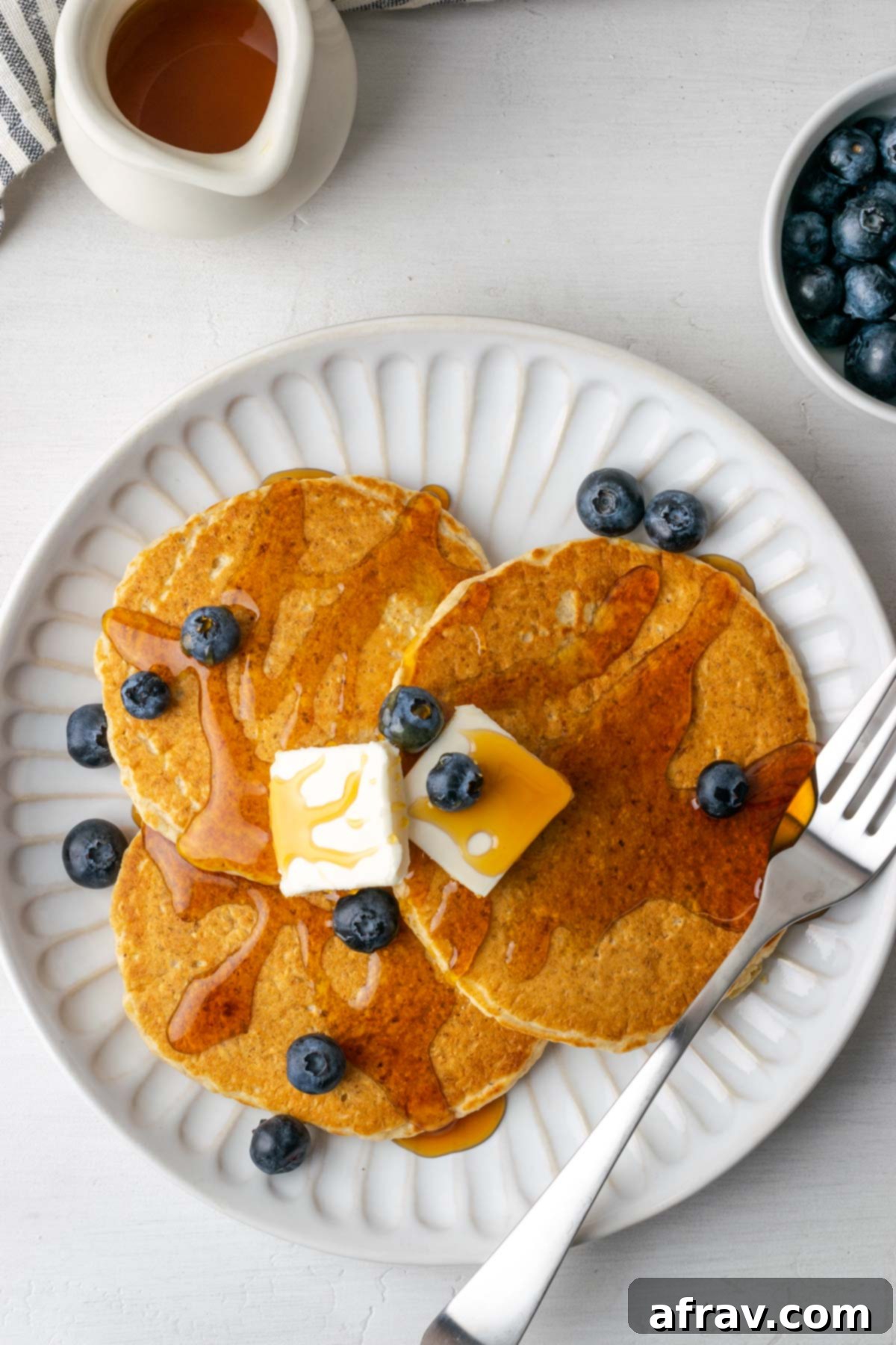 Three oat flour pancakes topped with butter, blueberries, and syrup on a round white plate.