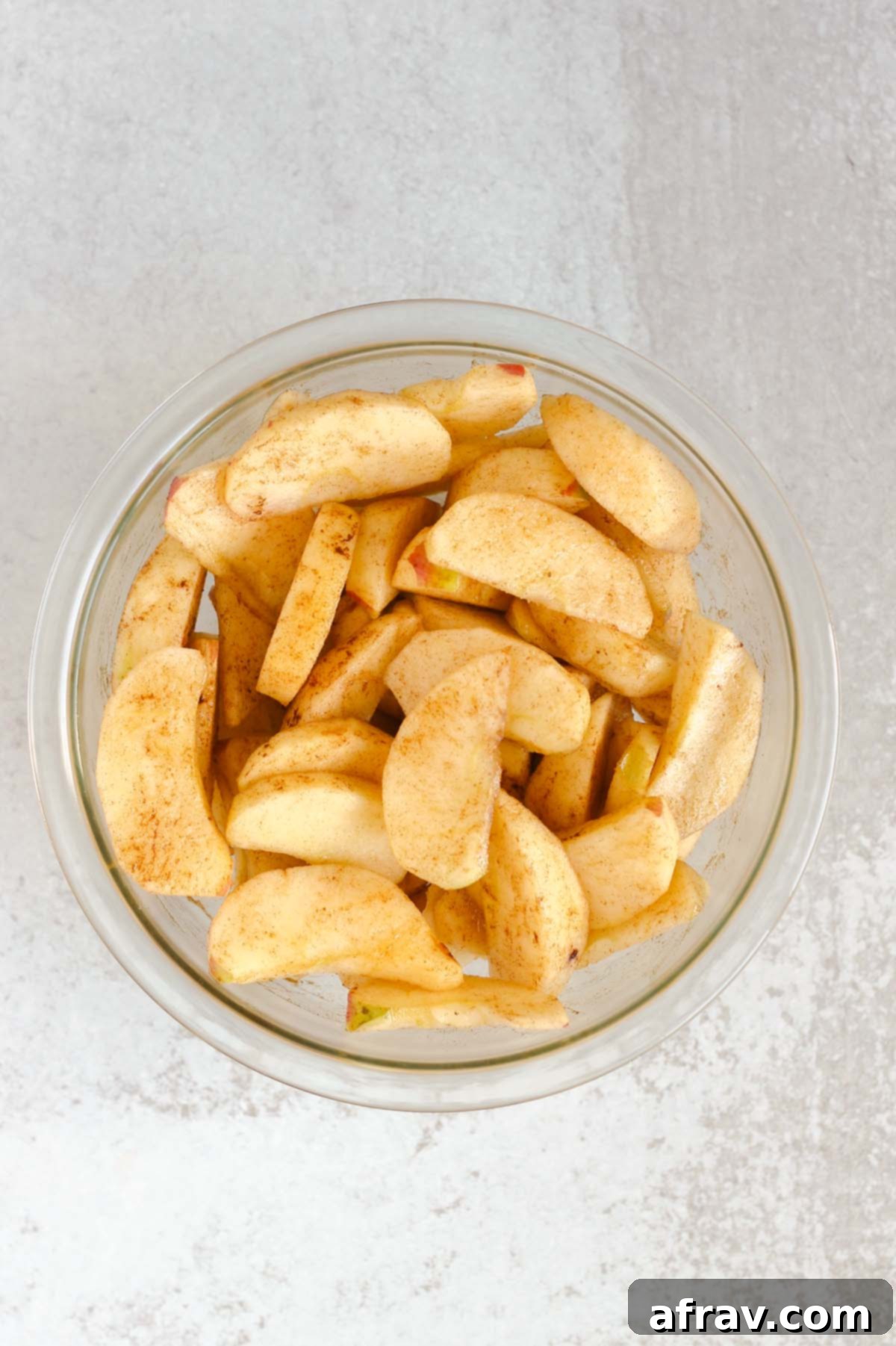 Apple slices mixed with avocado oil and cinnamon in a clear glass mixing bowl, ensuring even coating.