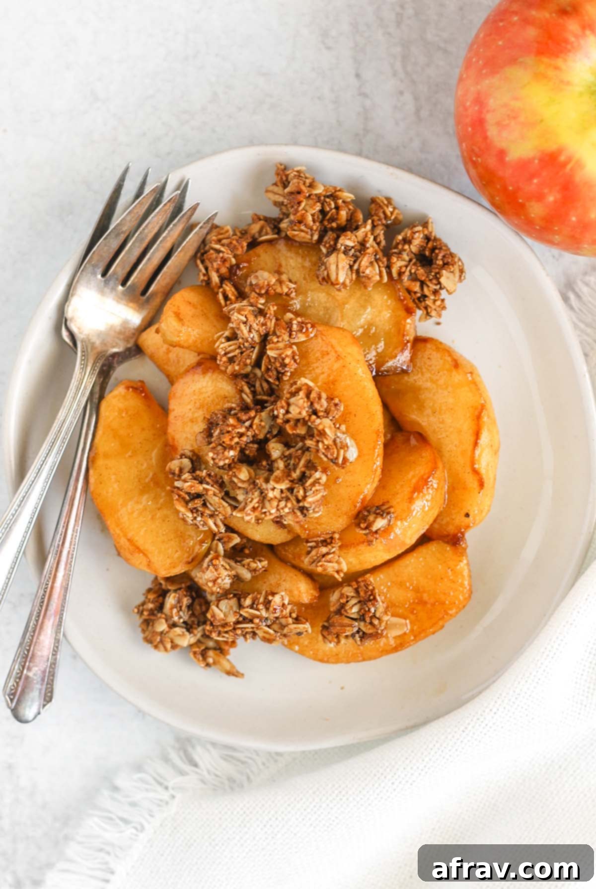 A serving of air fried apples topped with crunchy granola on a white plate, accompanied by vintage forks and a whole apple.