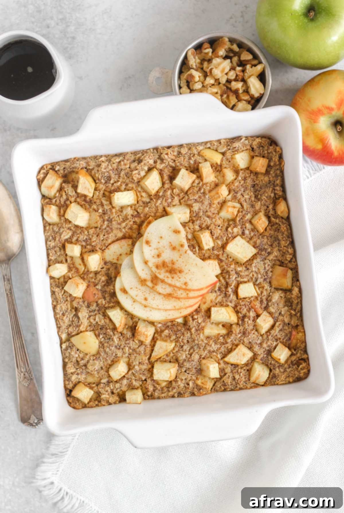 Apple cinnamon baked oatmeal topped with apple sliced and cinnamon in a white square baking dish with apples and syrup off to the side.